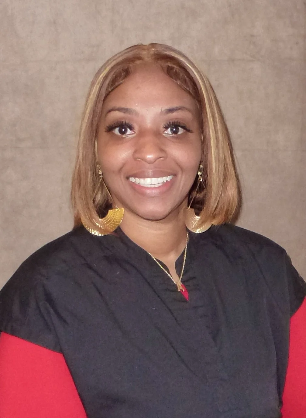 A woman smiling, wearing a black top with a red shirt underneath, gold hoop earrings, and a gold necklace.