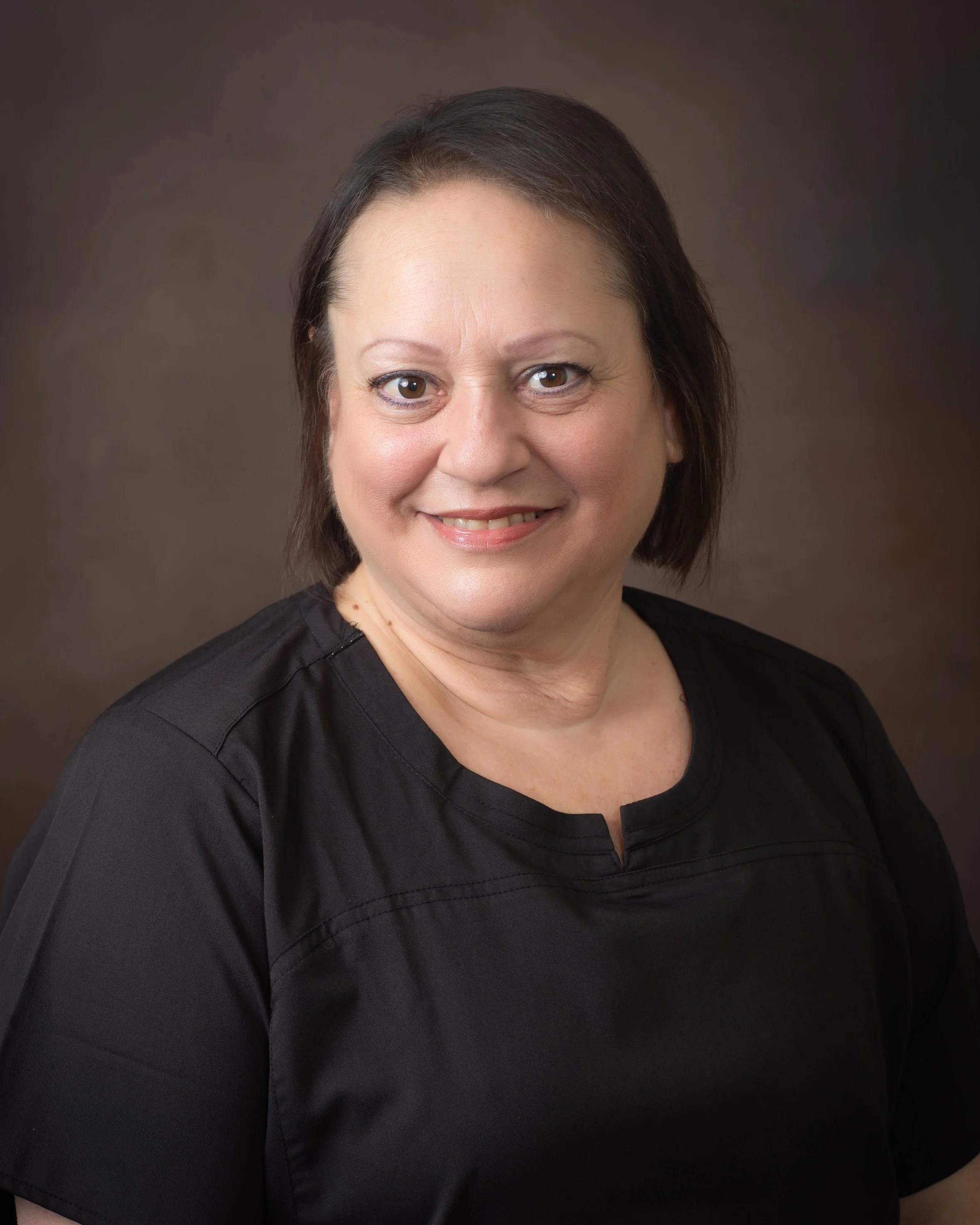Portrait of a woman smiling with short dark hair, wearing a black top, against a plain dark background.