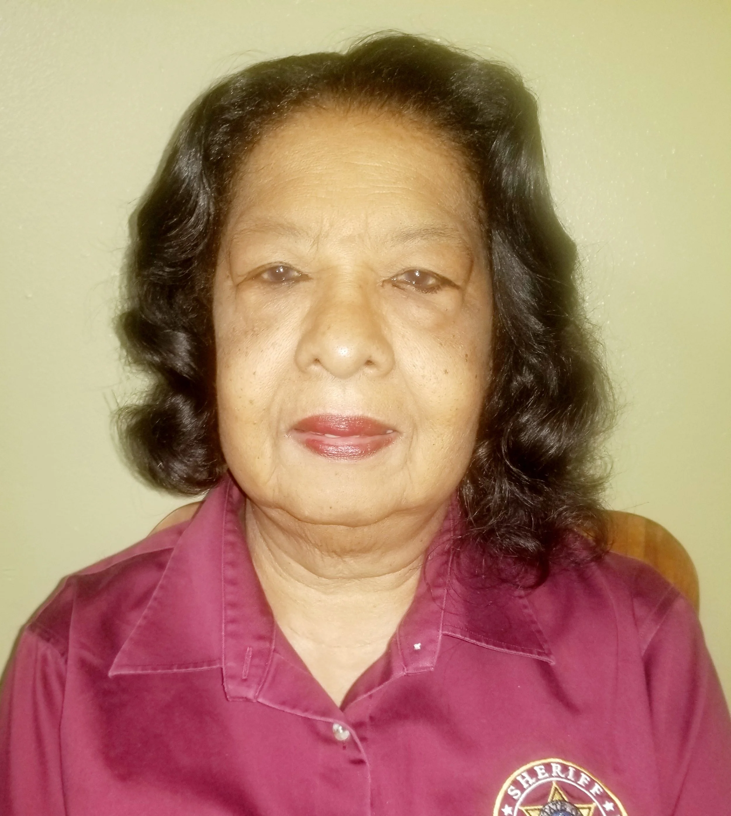 A person wearing a maroon shirt with a sheriff logo on it, posing for a portrait against a light-colored wall.