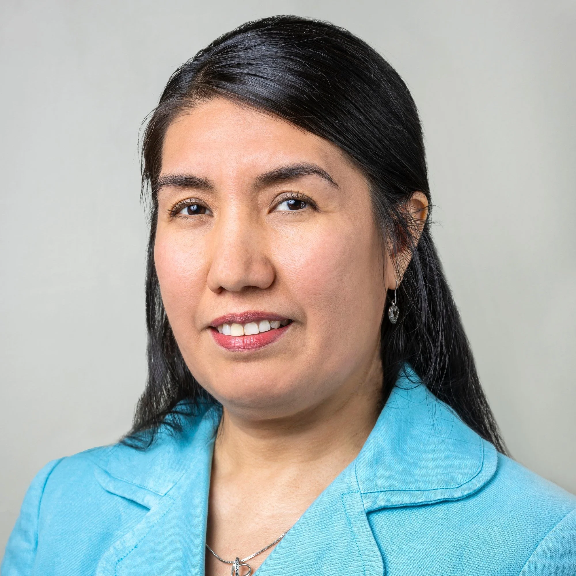 A woman wearing a light blue blazer with long dark hair, smiling, and facing the camera. Her expression is friendly and she has earrings and a necklace.