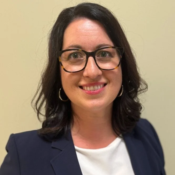 Smiling woman with glasses and dark hair wearing a navy blue blazer and white top against a neutral background.