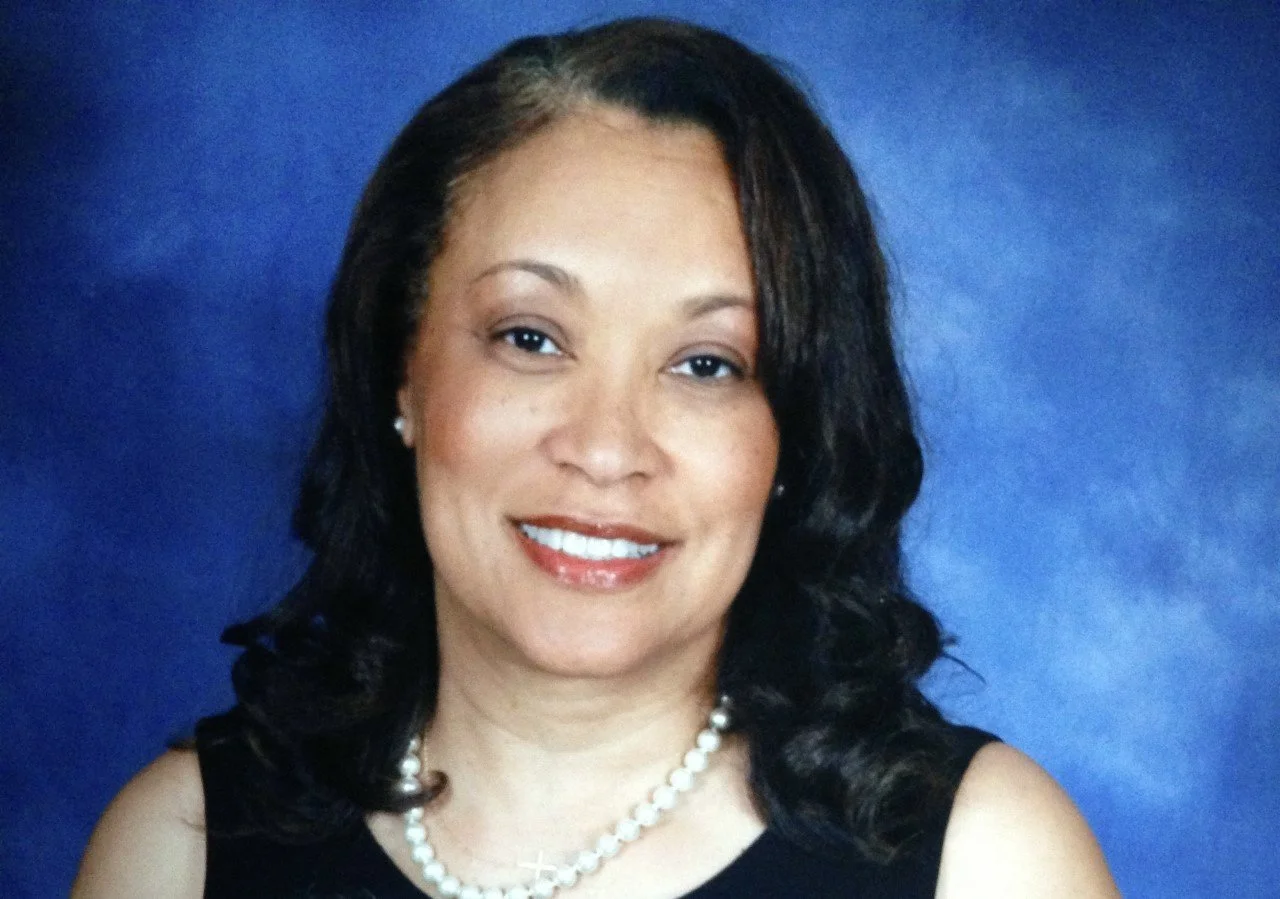 Portrait of a smiling woman with curly hair, wearing a pearl necklace and black top, against a blue background.
