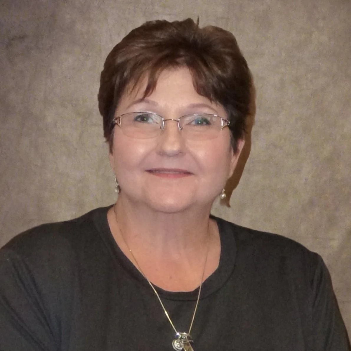 Middle-aged woman with short hair, wearing eyeglasses and a dark shirt, smiling against a neutral background.