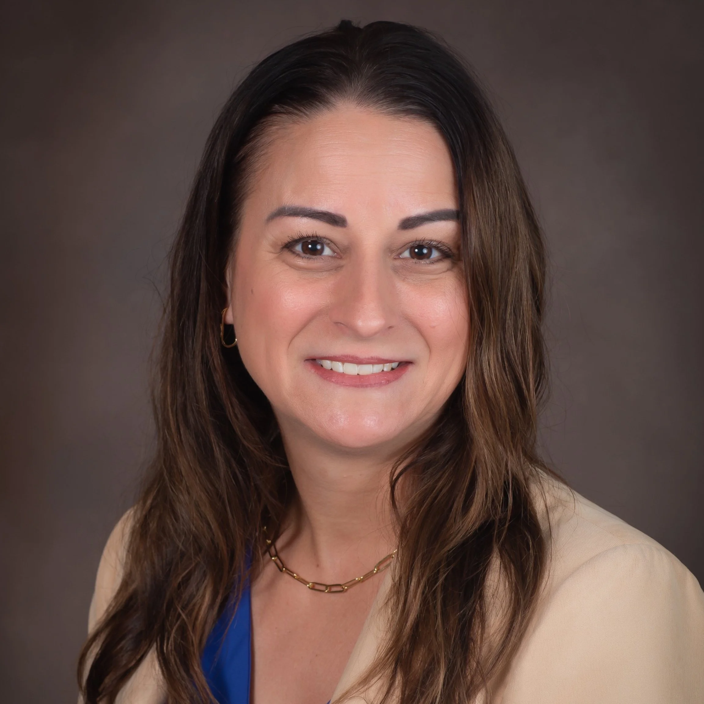 A woman with long brown hair, wearing a beige blazer, blue top, and a thin gold necklace, smiling against a plain brown background.