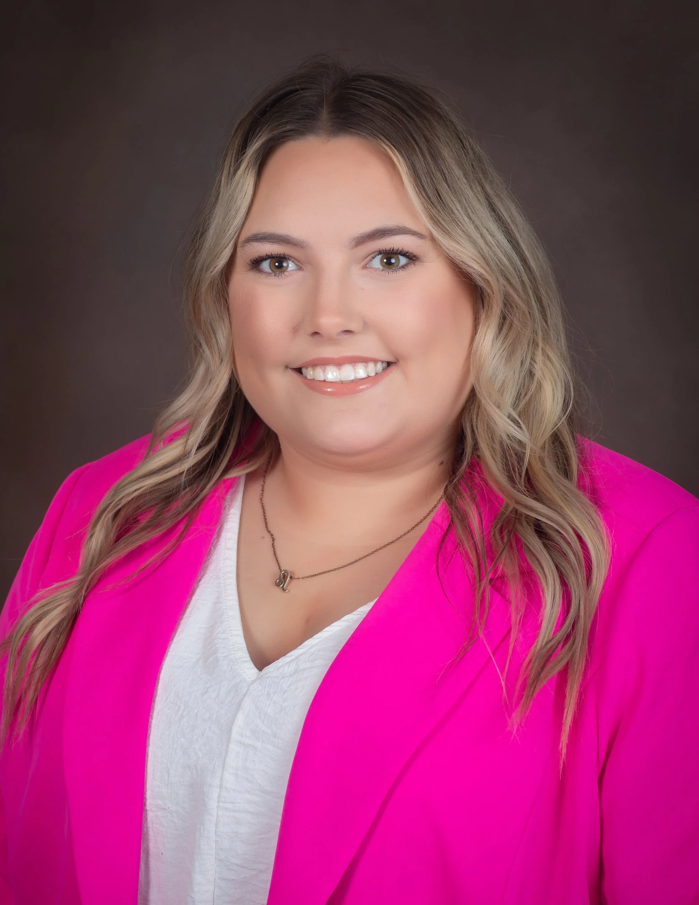 Woman with blonde hair smiling in a pink blazer and white top against a dark background.