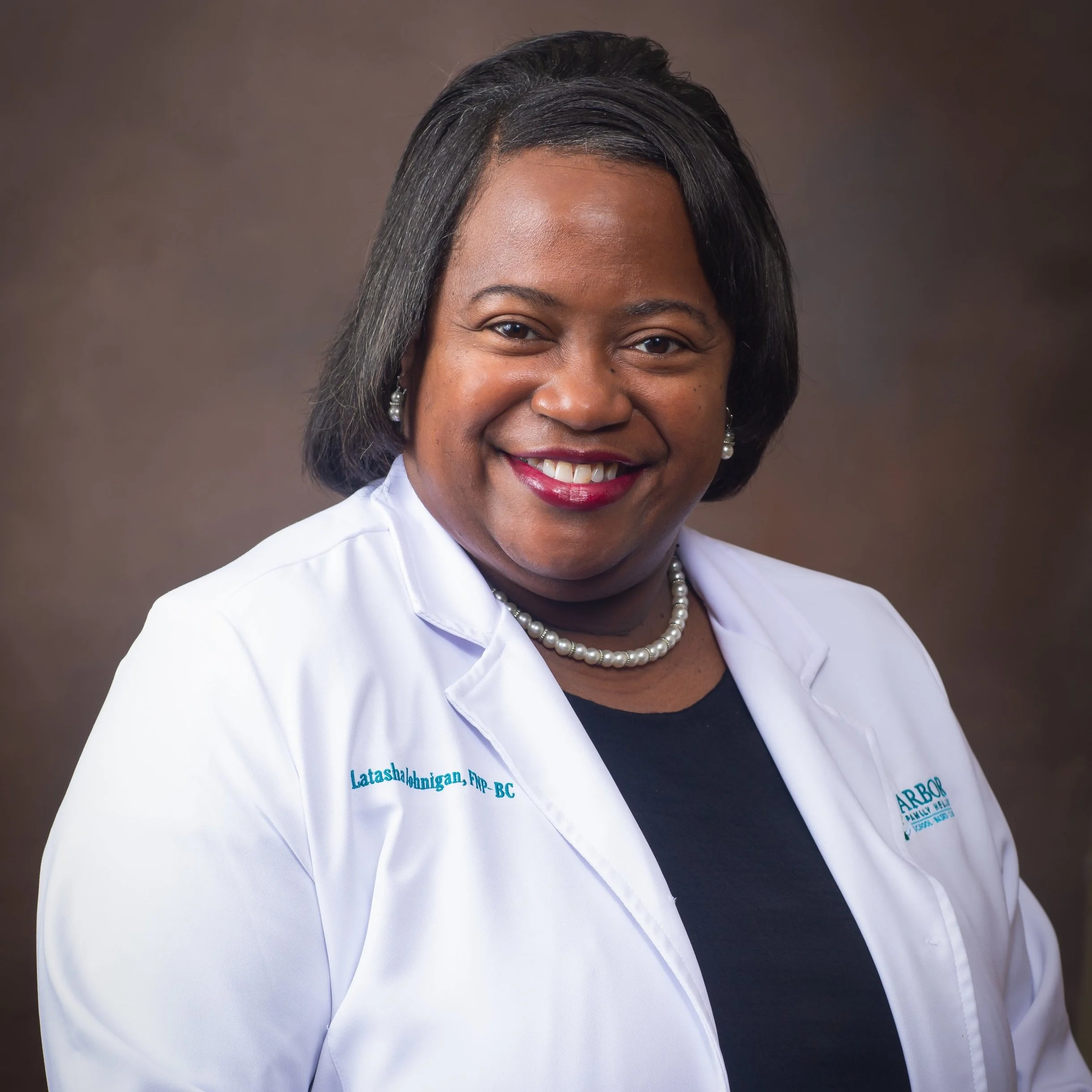 Professional woman in white medical coat with a pearl necklace, smiling against a neutral backdrop.