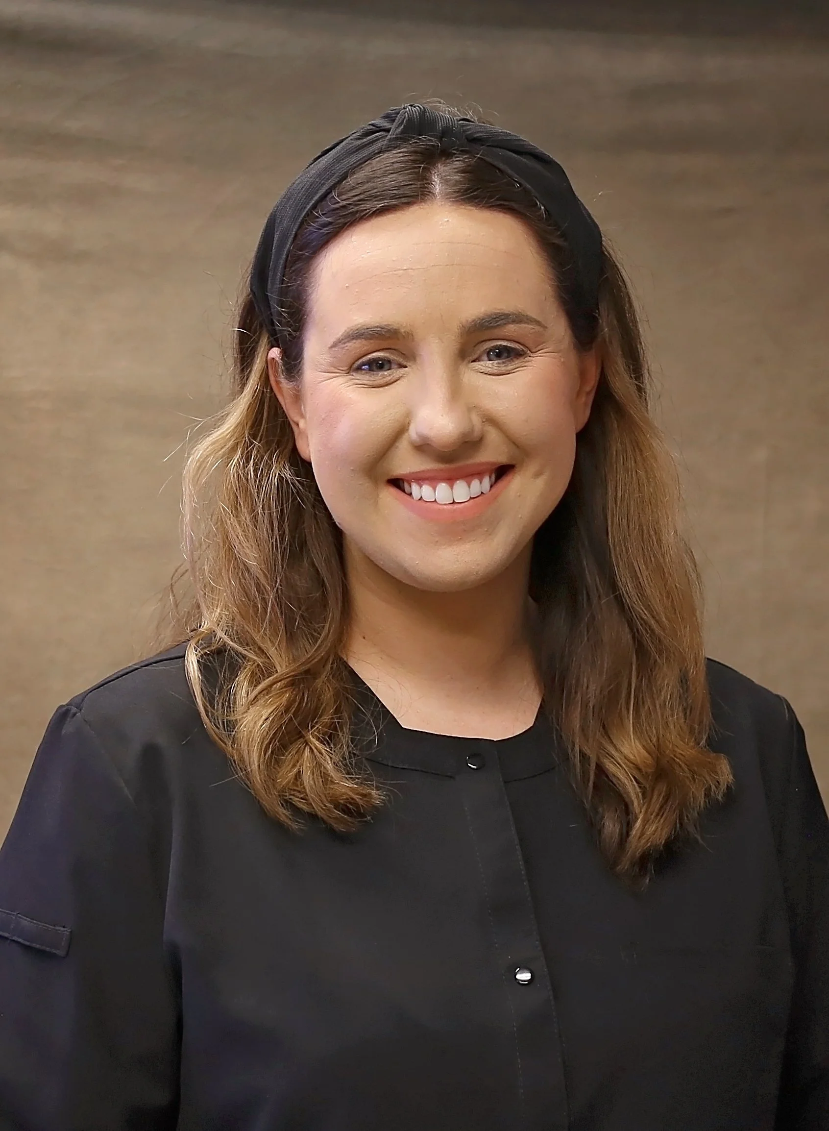 A smiling woman with shoulder-length light brown hair, wearing a black headband and a black shirt, standing against a plain brown background.