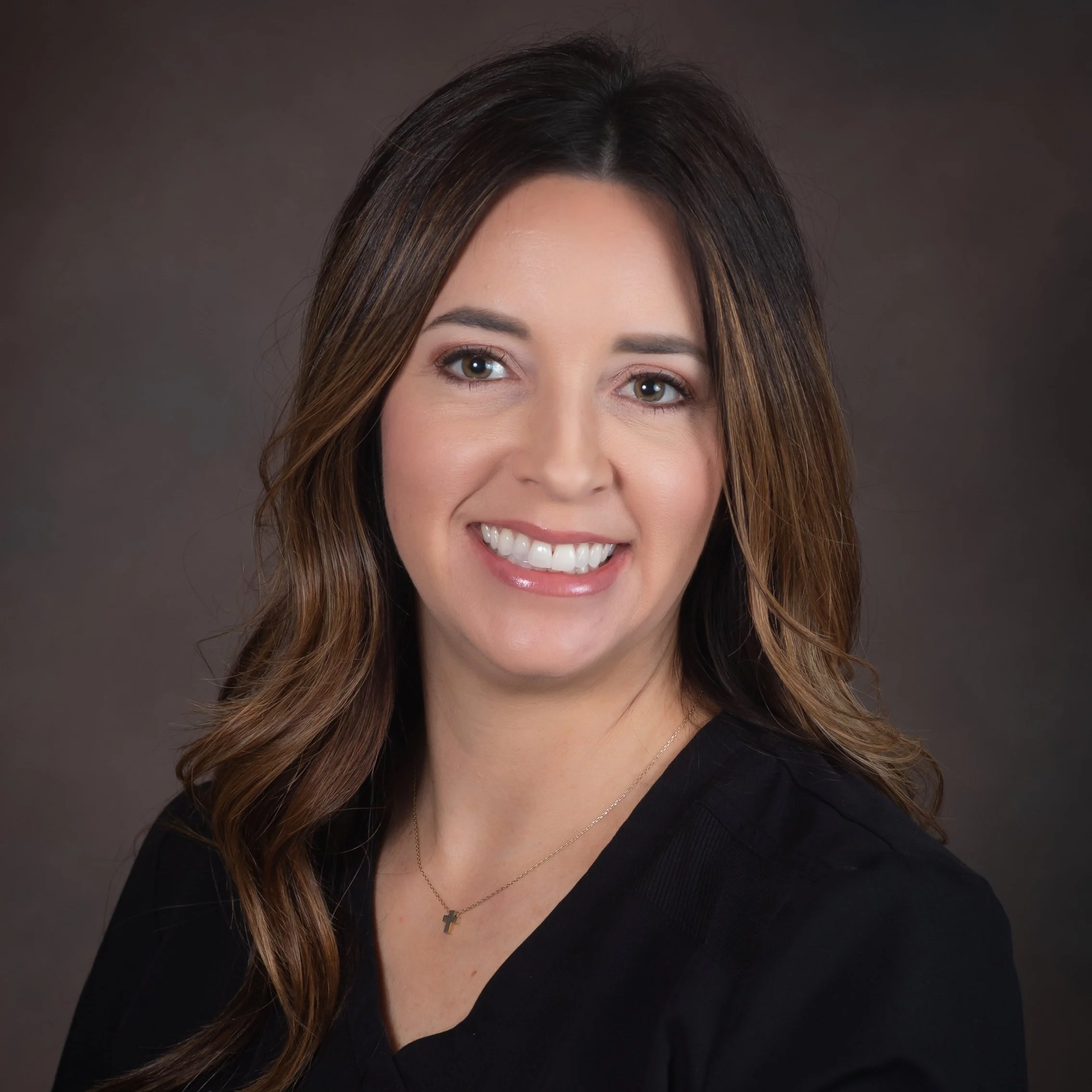 Portrait of a smiling woman with long brown hair wearing a black top and a necklace with a small cross pendant, set against a neutral brown background.