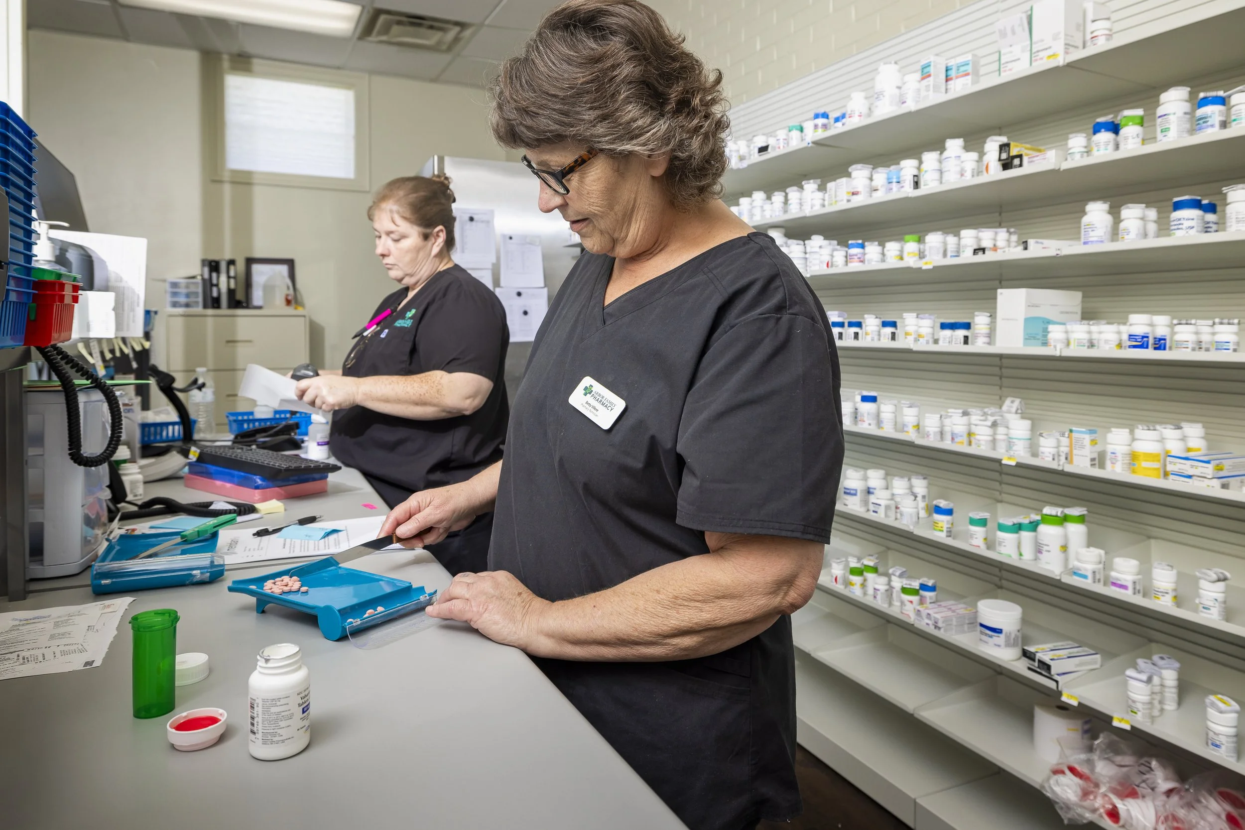 Two pharmacists sorting medication behind a counter in a pharmacy, with shelves of various medicine bottles in the background.