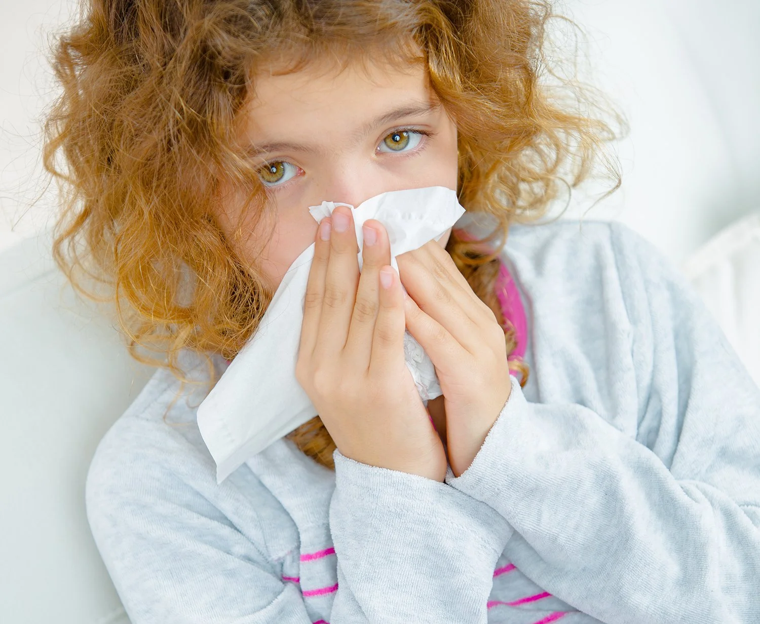 A young girl with curly red hair and green eyes holding a tissue to her nose, possibly sneezing or blowing her nose, wearing a light gray long-sleeve shirt with pink stripes.