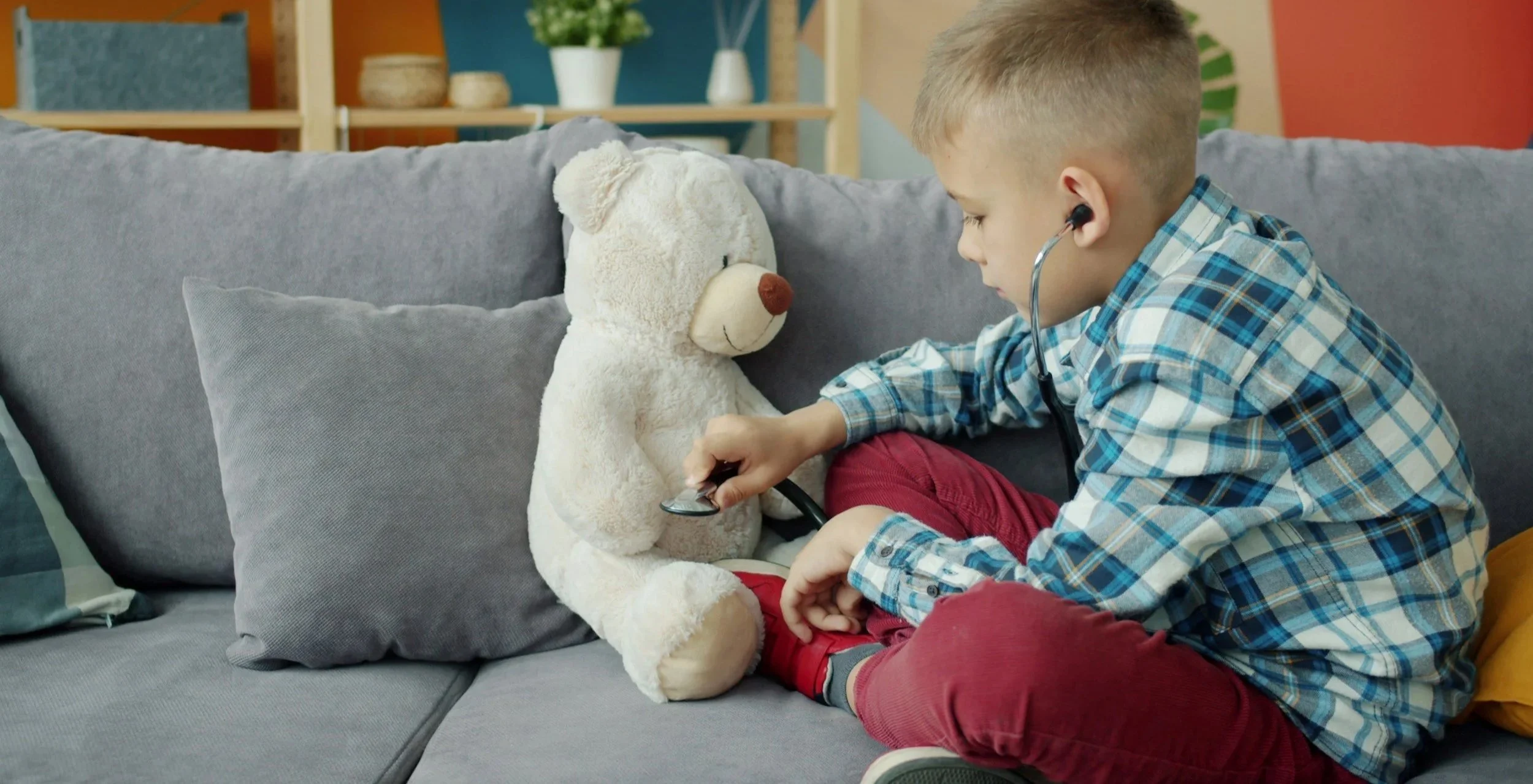 A young boy using a stethoscope to listen to a teddy bear's chest on a couch.