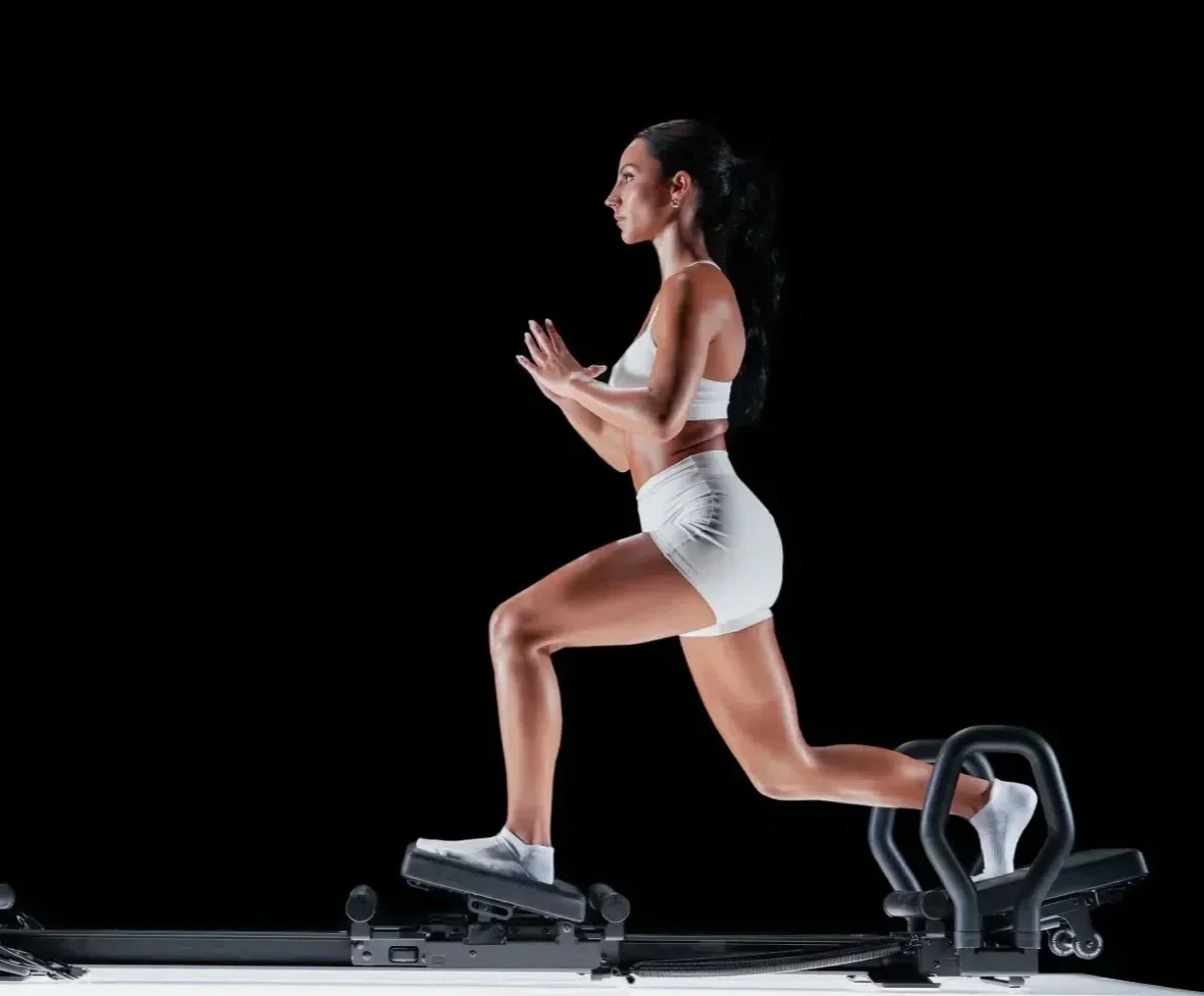A woman performing a lagree workout on a mini pro machine against a black background, wearing white sportswear.