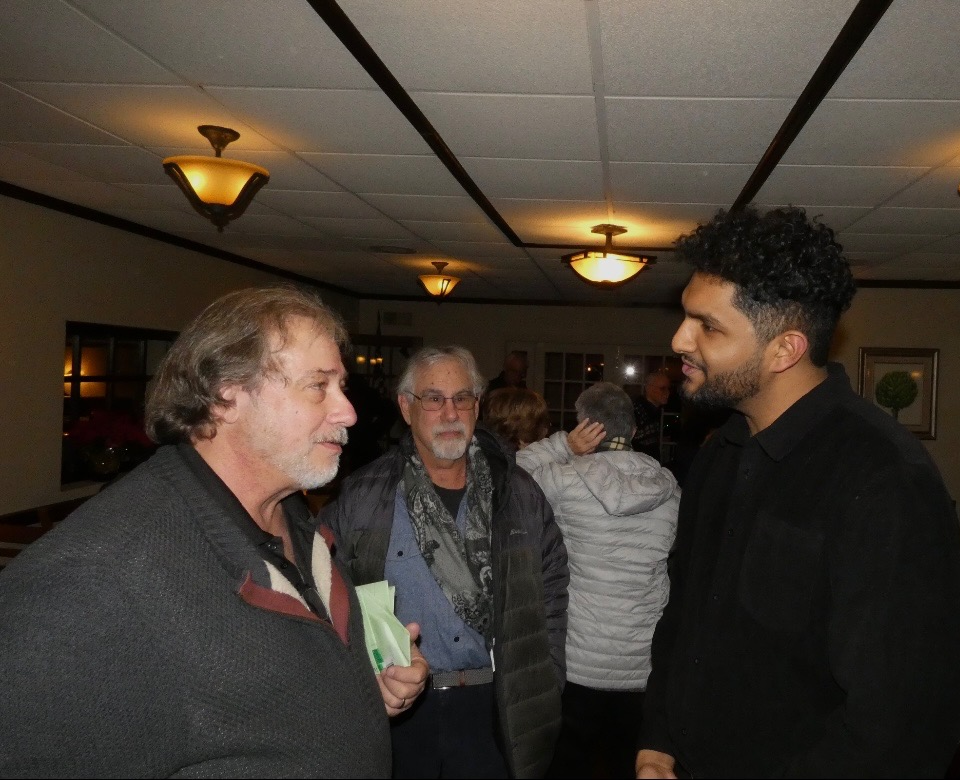 Farzeen Bham and two other men engaged in a conversation in a warmly lit indoor setting, with more people in the background.