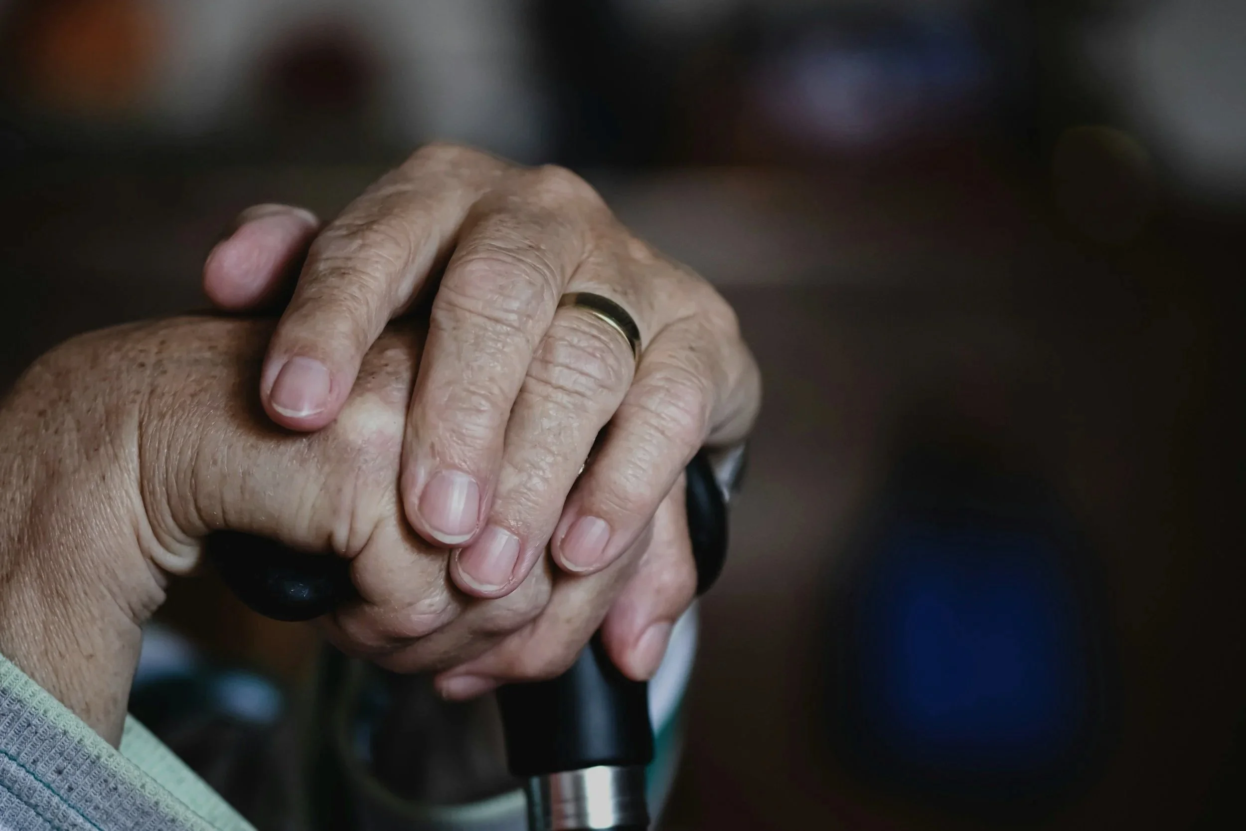 Close-up of two elderly hands clasped together, showing signs of age, with one hand wearing a wedding band. The background is blurred.