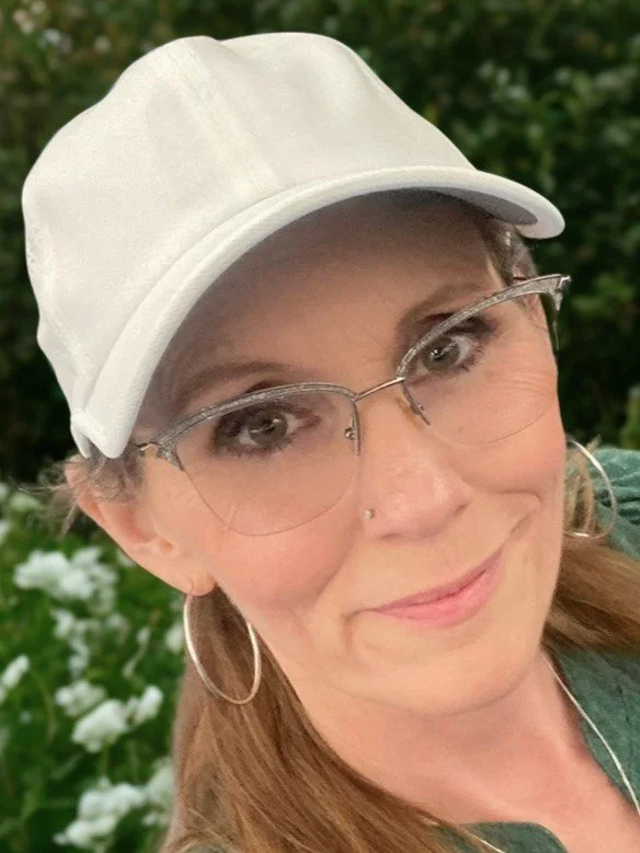A woman smiling outdoors, wearing a white baseball cap, glasses, hoop earrings, and a green shirt.