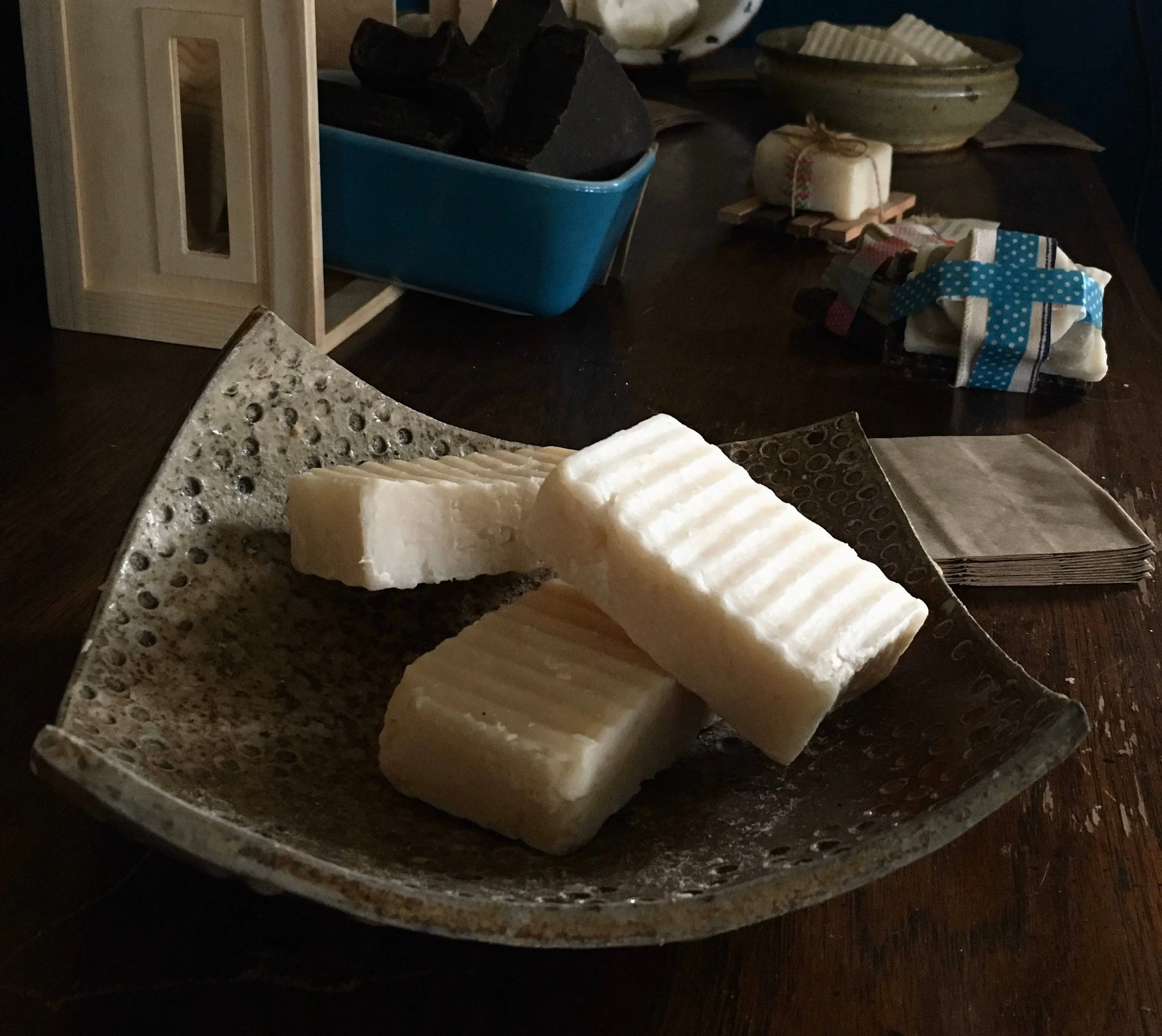 Four bars of handmade soap on a decorative ceramic dish on a wooden table with soap-making supplies and folded cloths in the background.