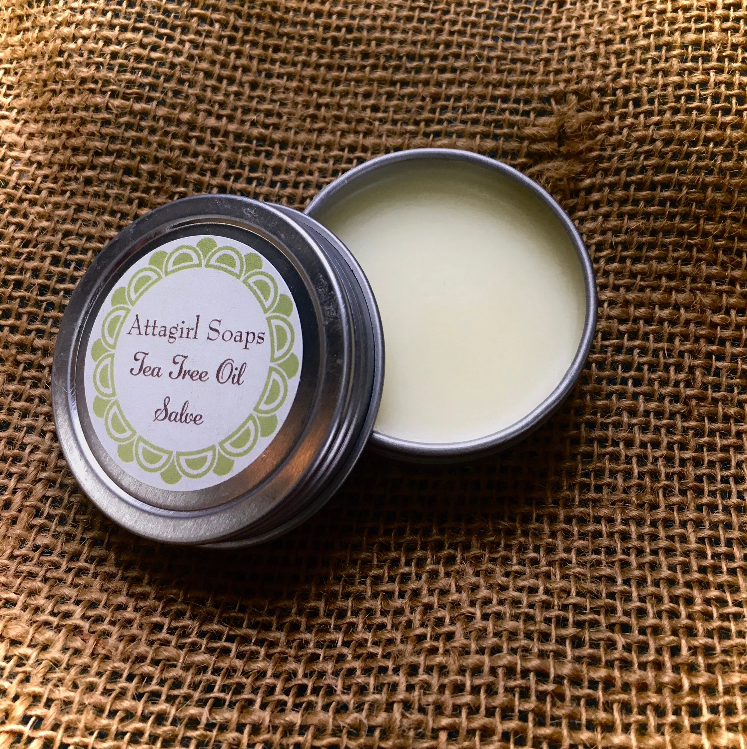 Open tin container of Attagirl Soaps Tea Tree Oil Salve on textured brown fabric background.