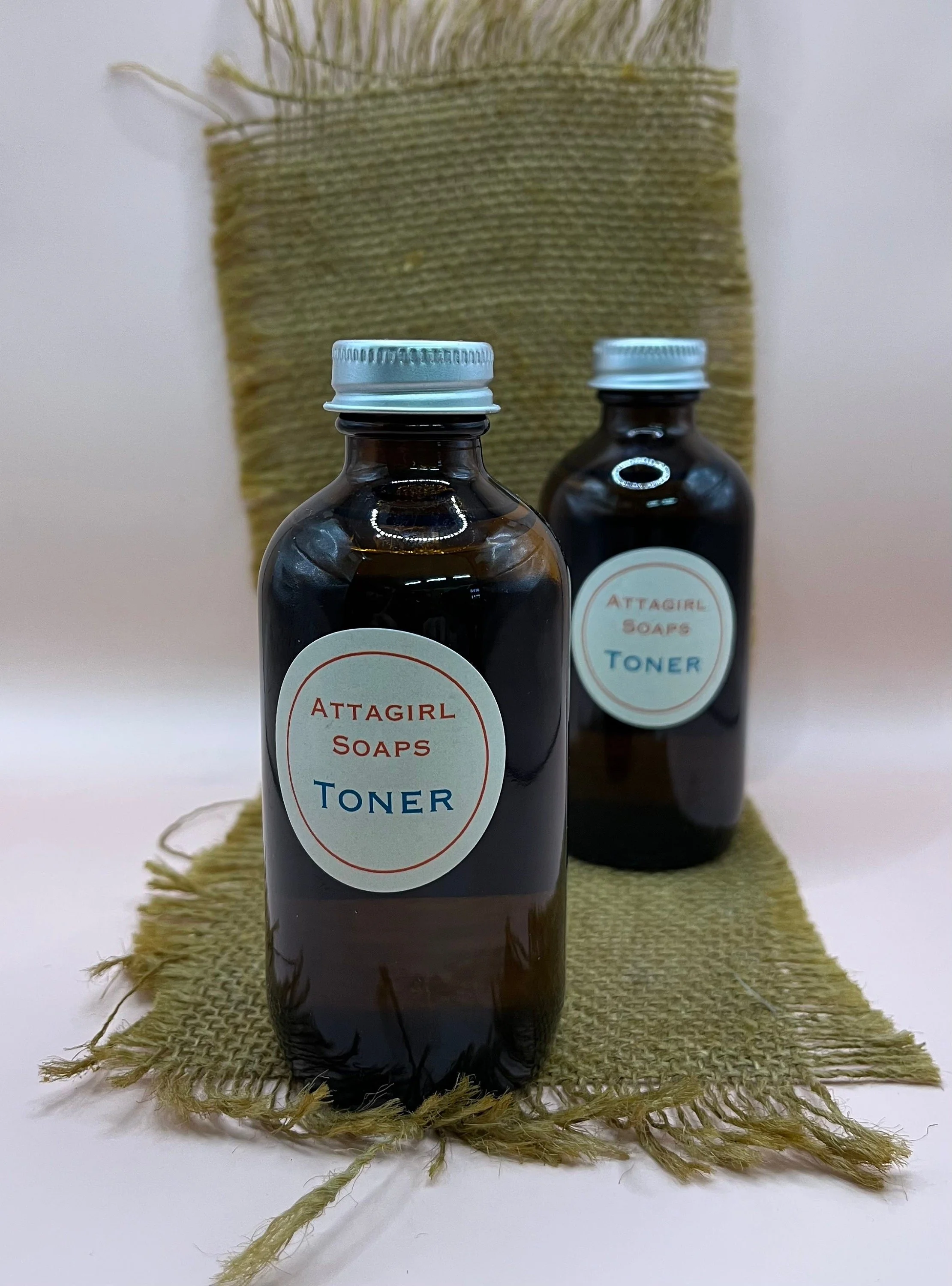 Two dark glass bottles labeled 'Attagirl Soaps Toner,' placed on a rough-textured, frayed piece of fabric, with a large piece of similar fabric behind them.