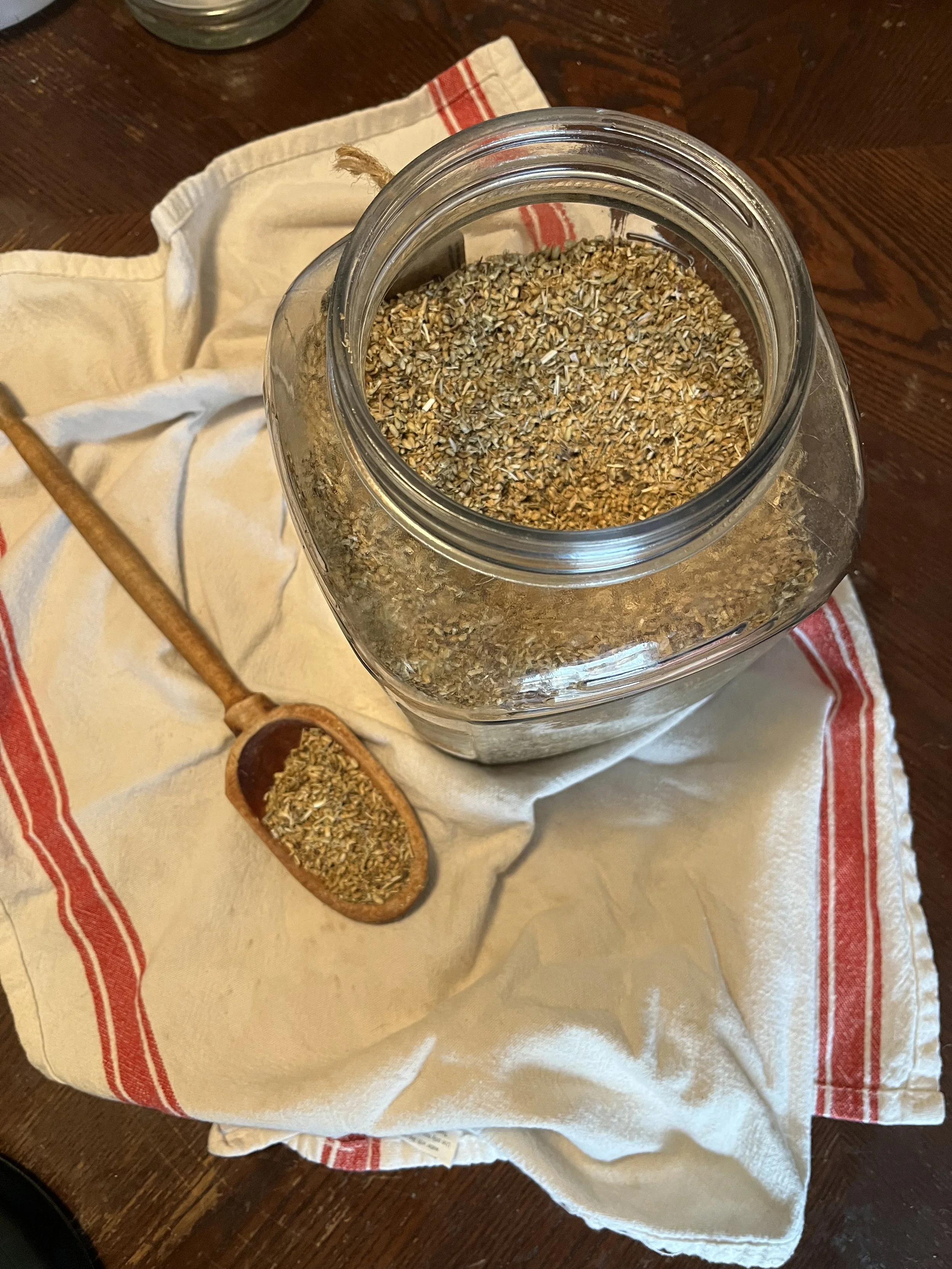 A glass jar filled with dried herbs on a cloth, with a wooden scoop containing similar herbs resting on the cloth.