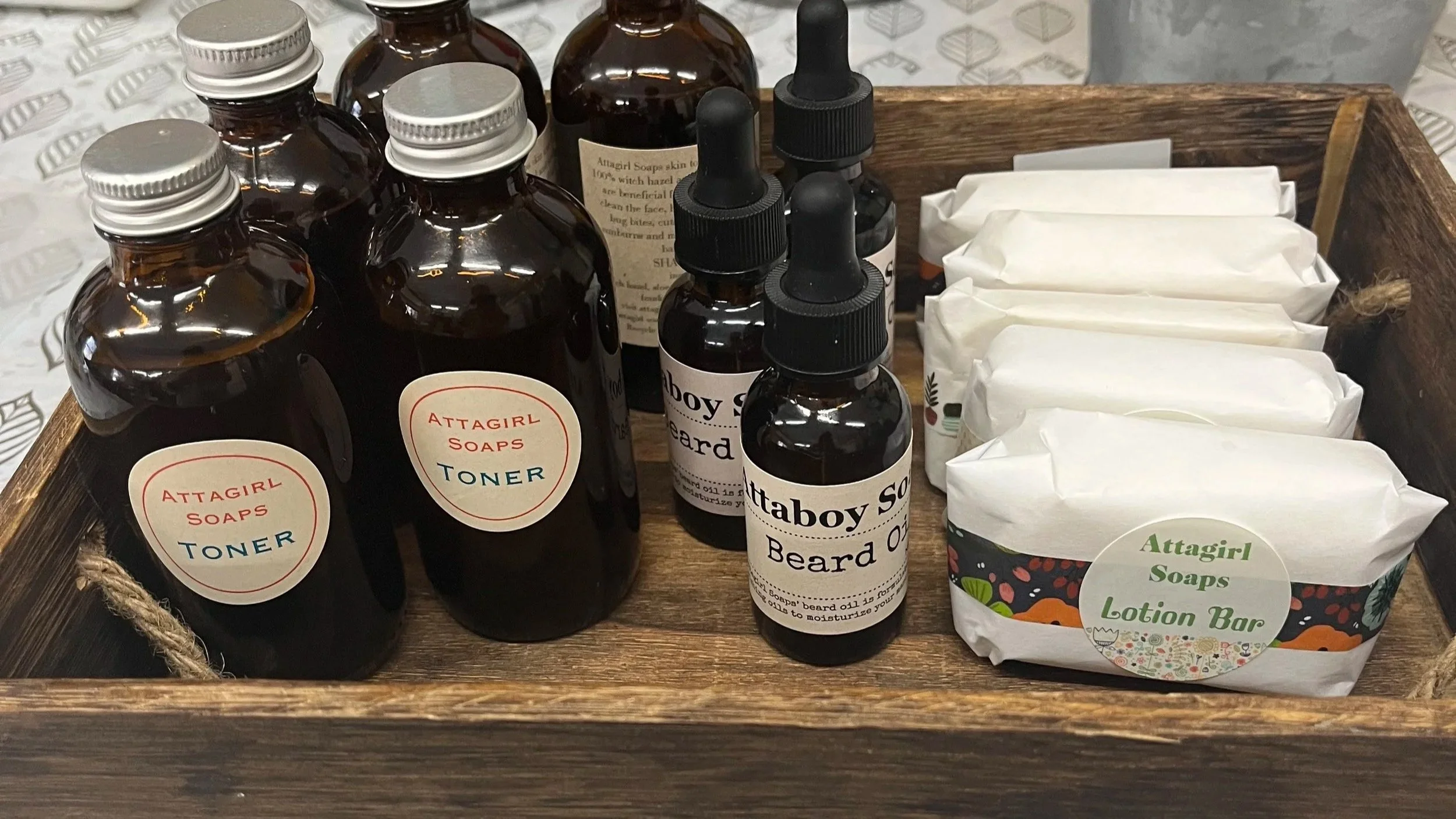 A wooden tray with bottles labeled 'Attagirl Soaps Toner,' 'Attagirl Soaps Beard Oil,' and 'Attagirl Soaps Lotion Bar,' along with wrapped soap bars.