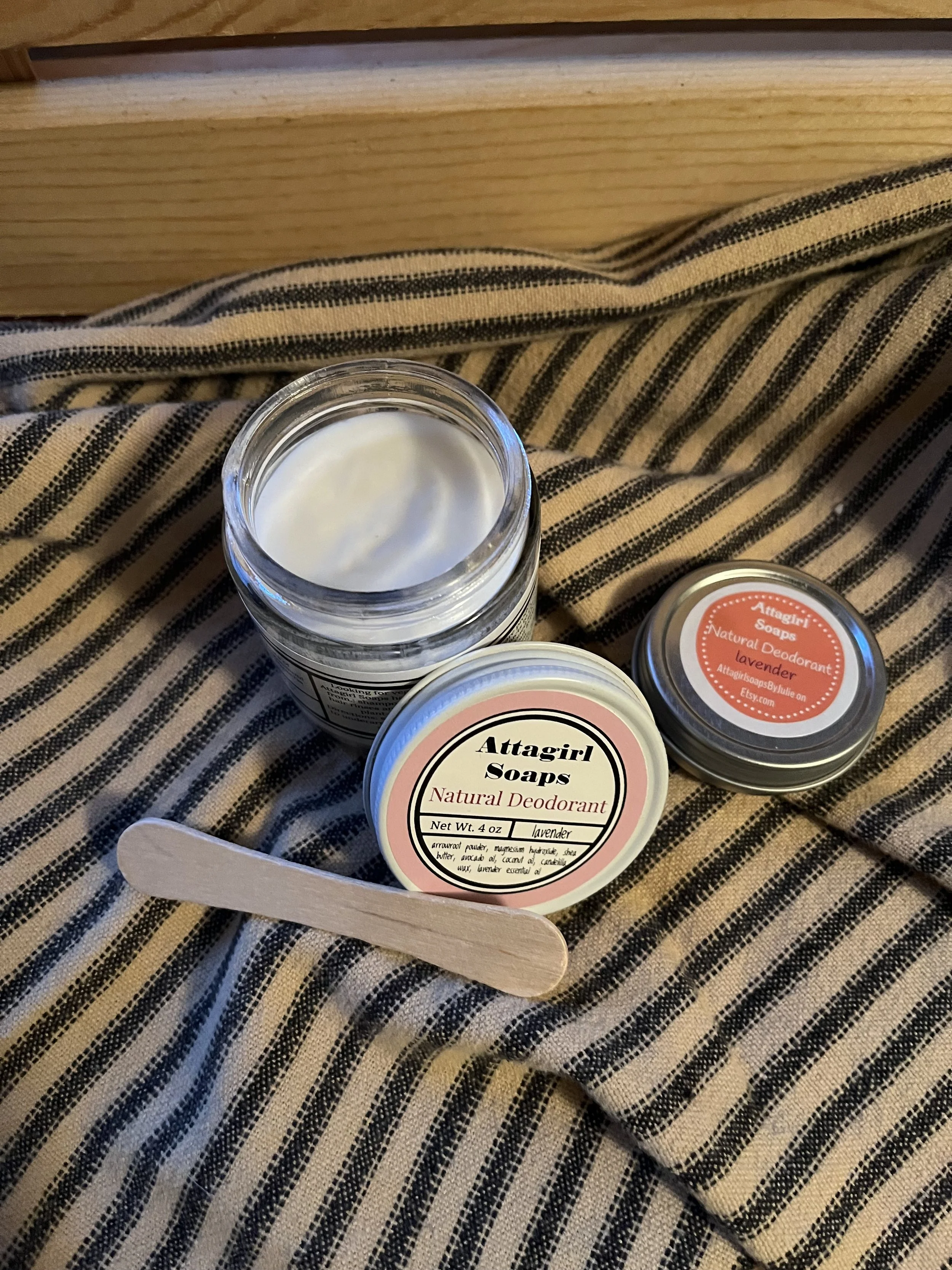 Open jar of Attagirl soap with white cream, a small container of Attagirl soap with lavender deodorant, and a wooden popsicle stick on black and beige striped fabric.
