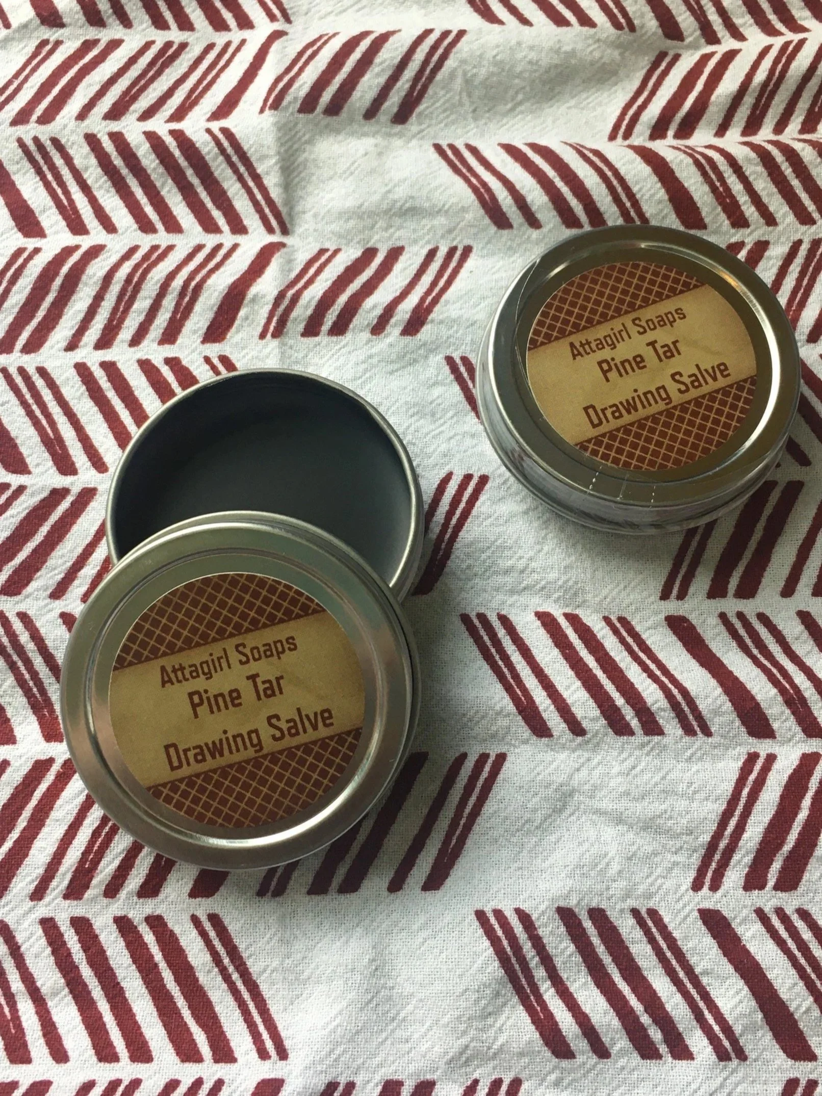 Two tins of Attagirl Soaps Pine Tar Drawing Salve on a patterned white and red cloth.