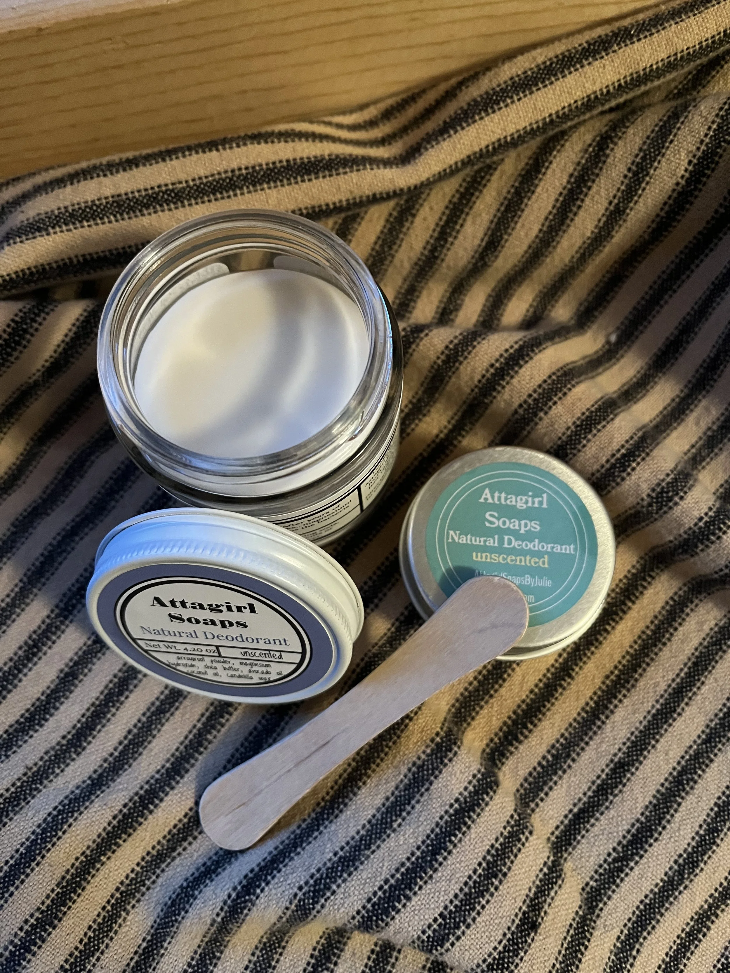 An open jar of natural deodorant cream, two small containers of Attagirl Soaps, and a wooden spatula on a striped fabric surface.