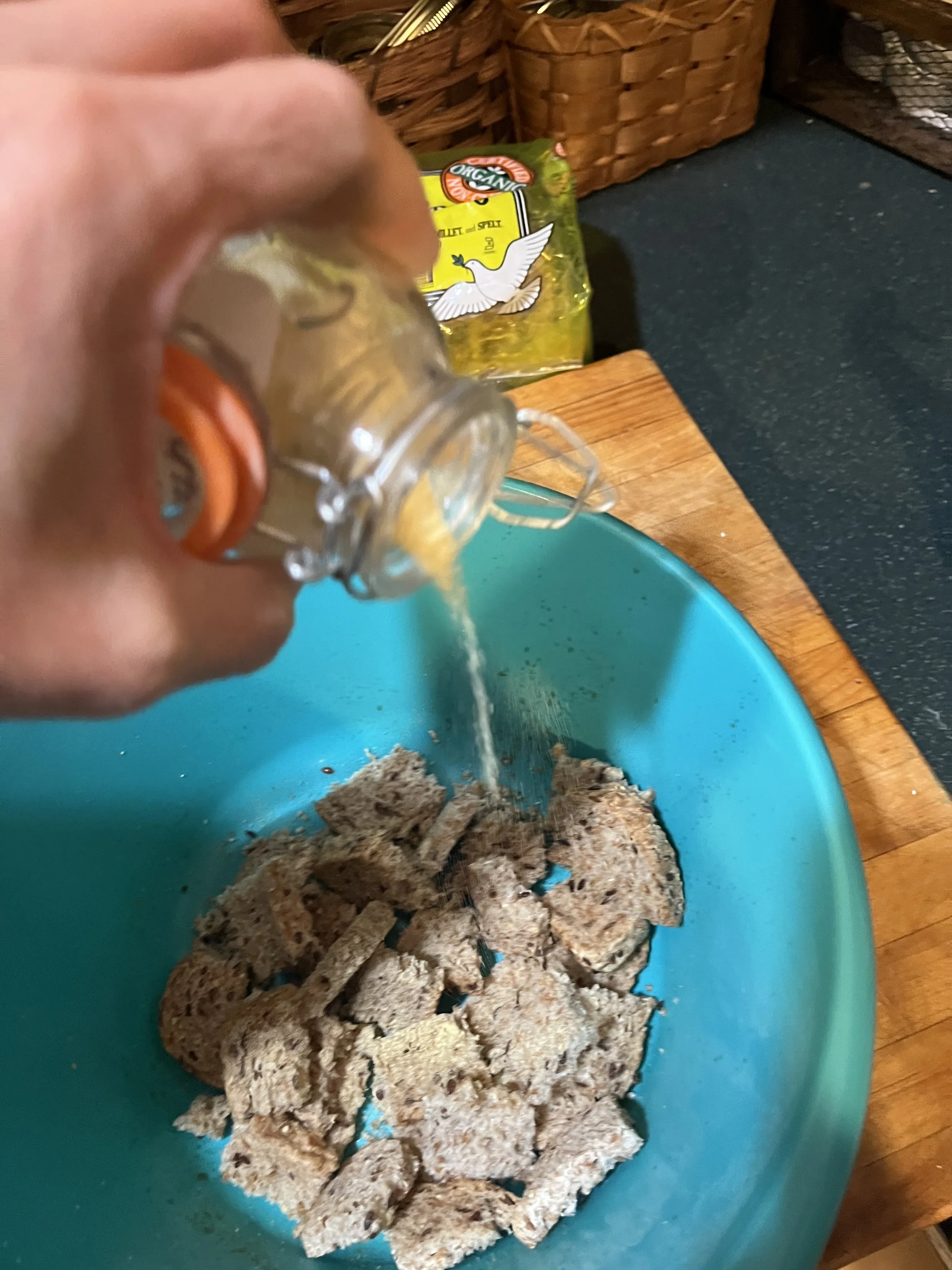 Pouring yellow liquid into a blue bowl filled with broken cereal pieces, on a wooden cutting board.