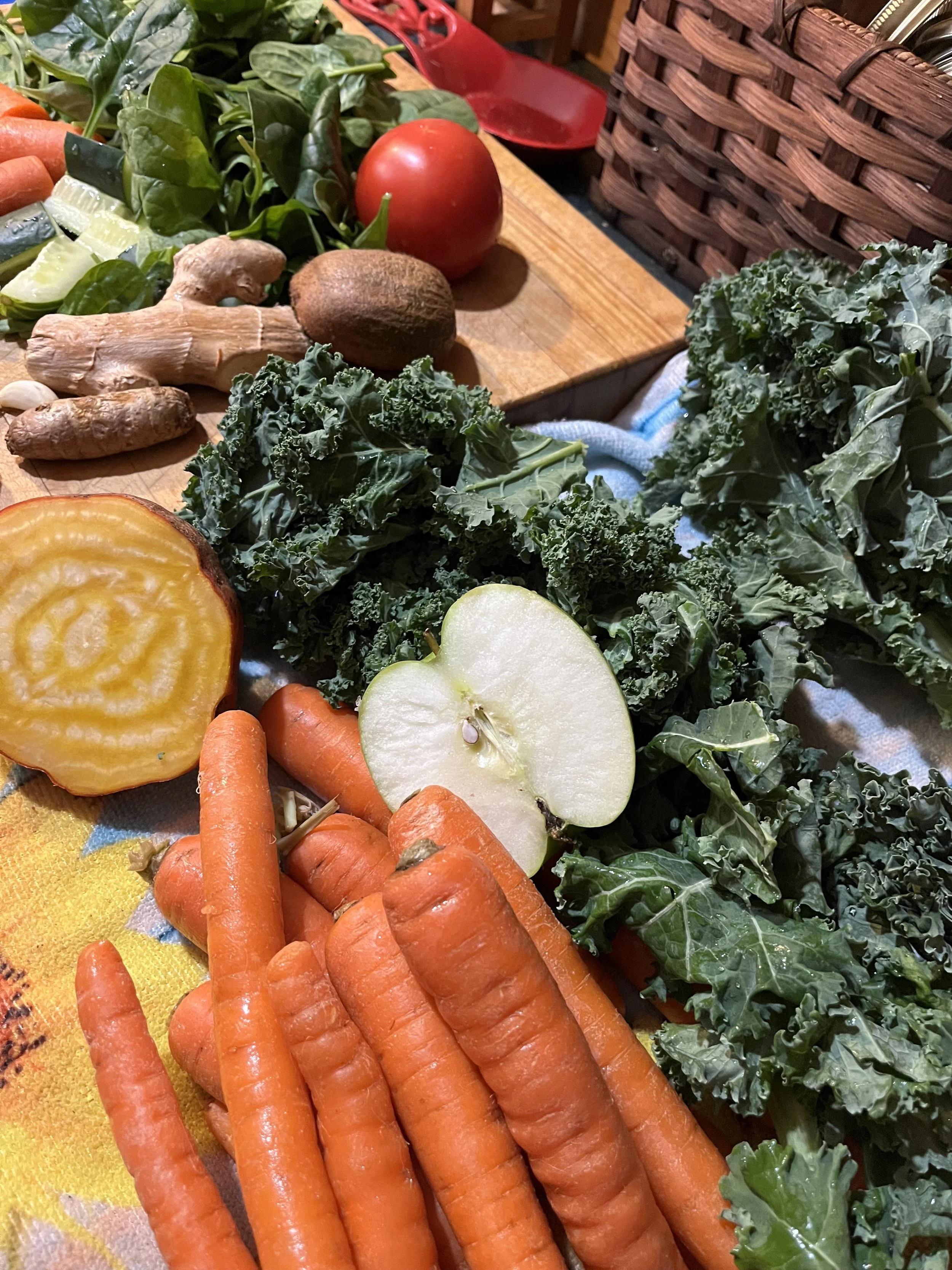 Fresh vegetables including carrots, leafy kale, a green apple, tomatoes, ginger, a mushroom, a lime, and a yellow squash on a kitchen counter.
