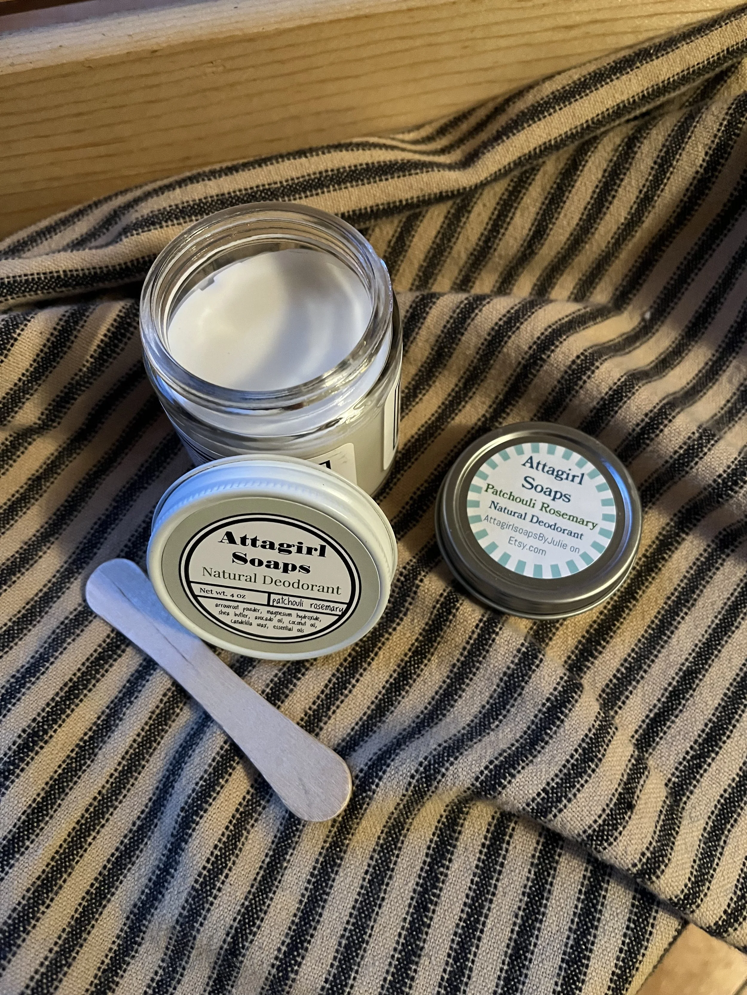 Open jar of Attagirl Soaps natural deodorant with a stick applicator and a separate container of Attagirl Soaps soap, both labeled with eucalyptus and patchouli rose scent, placed on a striped fabric surface.