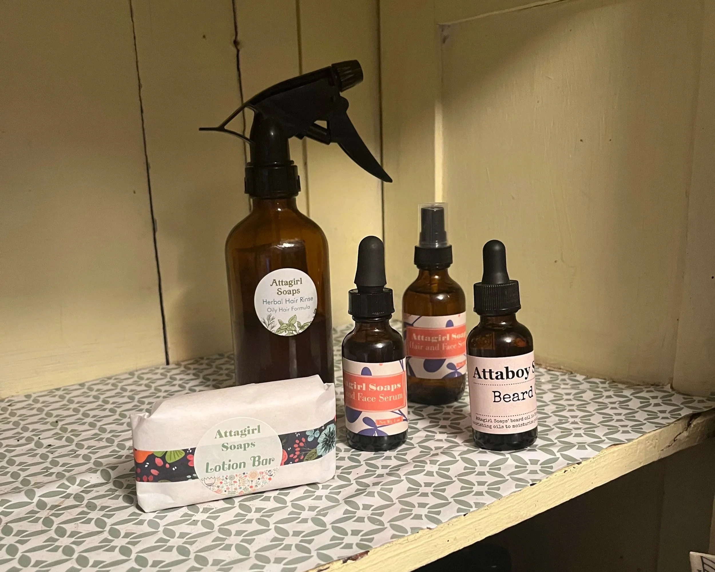 A collection of Attagirl Soaps products including a lotion bar, herbal hair rinse spray, face serum, beard oil, each with labels, displayed on a patterned shelf with a light-colored background.