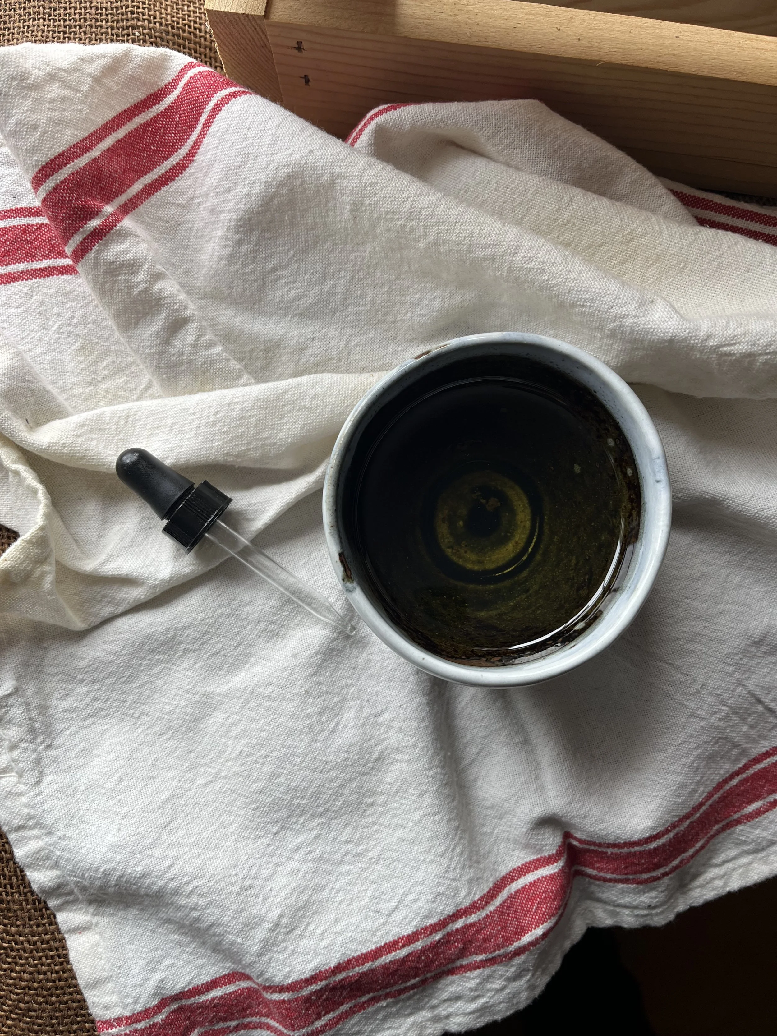 Top-down view of a ceramic cup filled with black coffee and a glass dropper with a black cap resting on white cloth with red stripe, placed on a textured surface near wooden furniture.