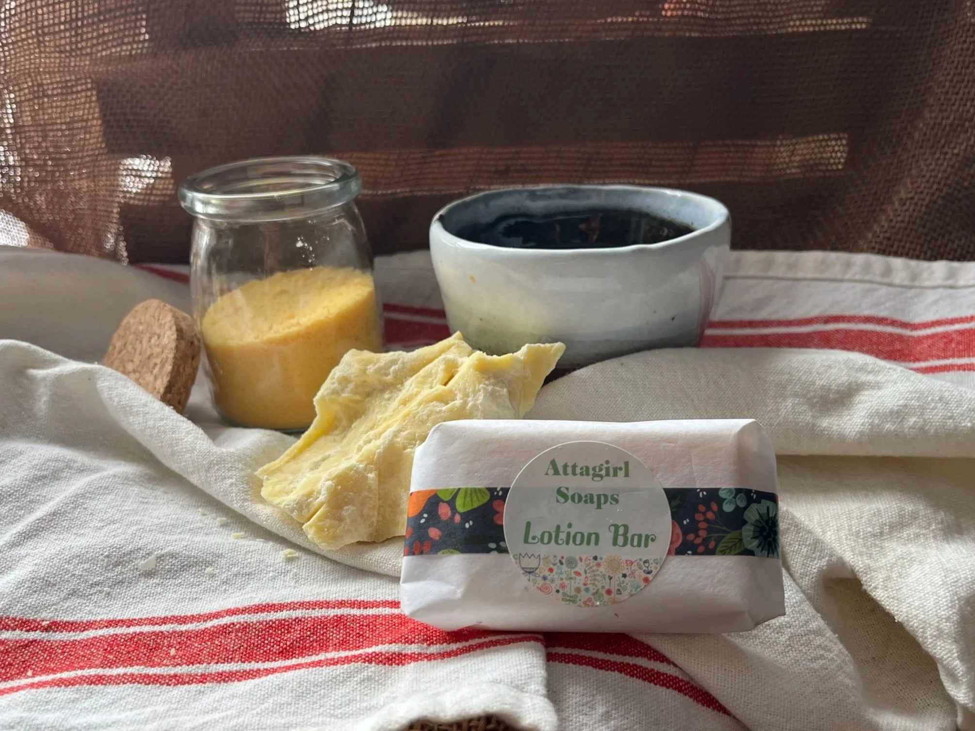 A still life of a setting on a cloth with a jar of candelila wax pellets, a piece of cocoa butter, a bowl of avocado oil and a solid bar of lotion labeled 'Attagirl Soaps Lotion Bar.'