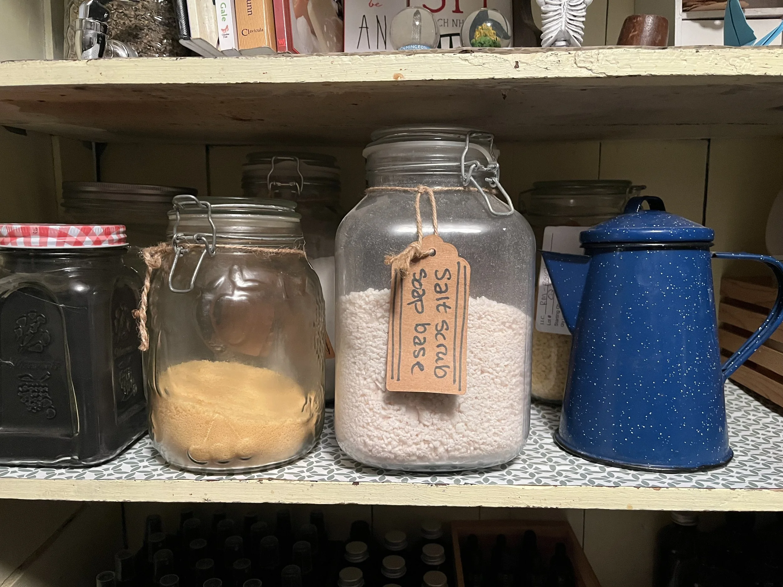 Glass jars containing dried yellow and white powders, with handwritten labels, on a kitchen shelf next to a blue speckled teapot.