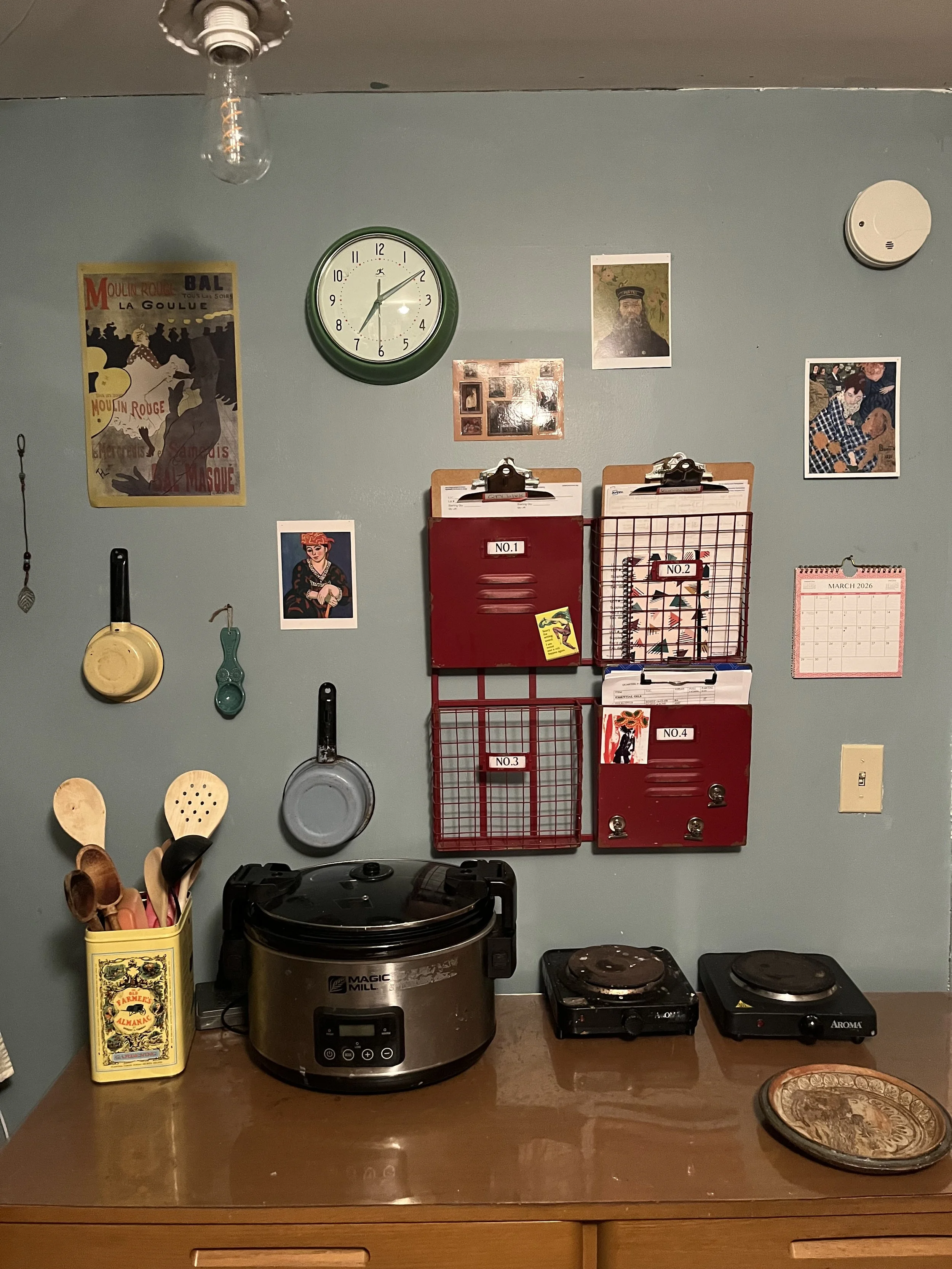 Kitchen countertop with utensils in a yellow container, a large crock pot, and a decorative plate. The wall behind features hanging utensils, vintage posters, framed pictures, a clock, a calendar, and several red wire baskets with labels.
