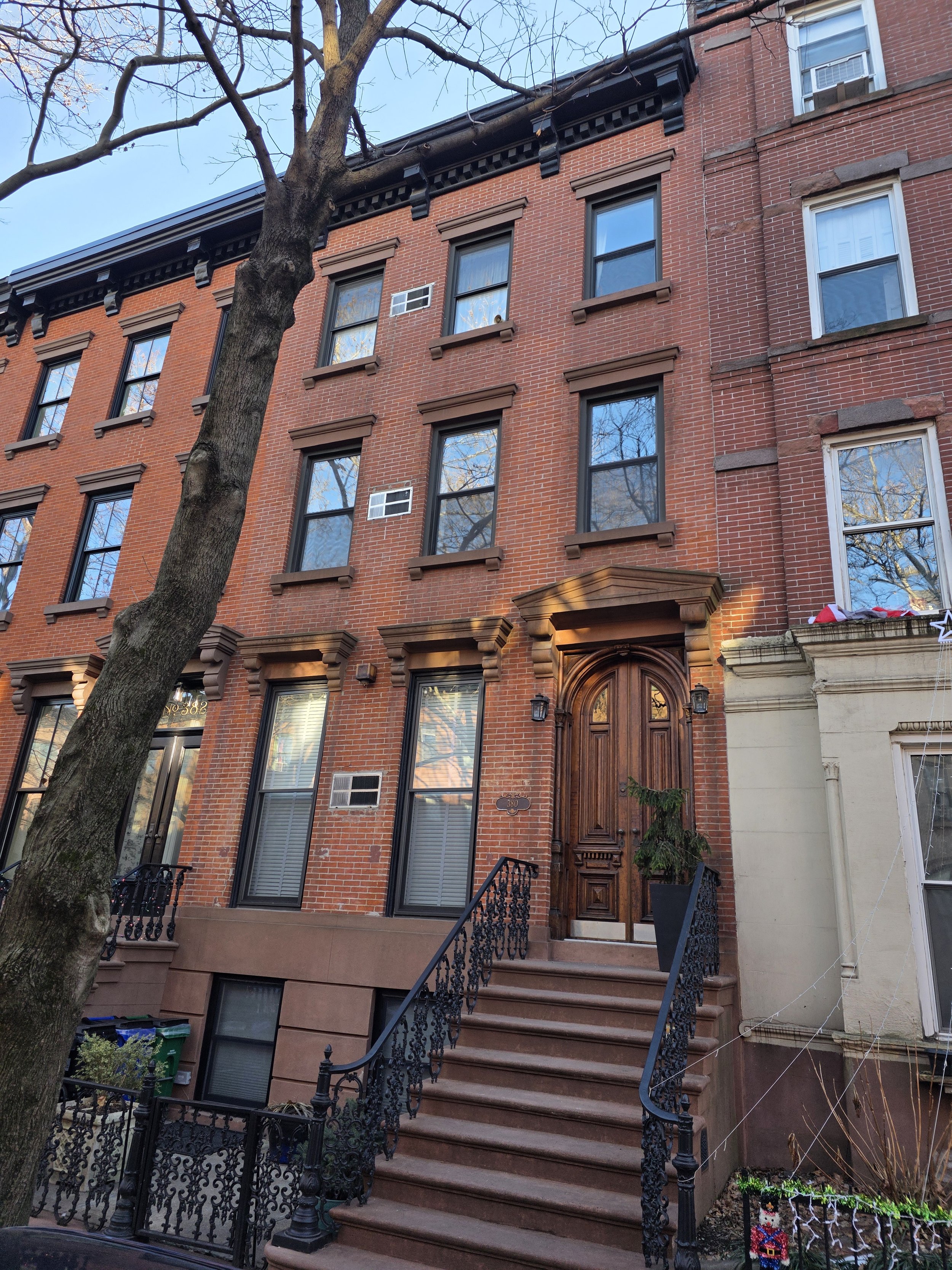 A red brick city townhouse with black window frames, a dark wood front door, and ornamental black iron railings on the staircase leading to the entrance. A tree is in front of the building, and a potted plant is on the steps near the door.
