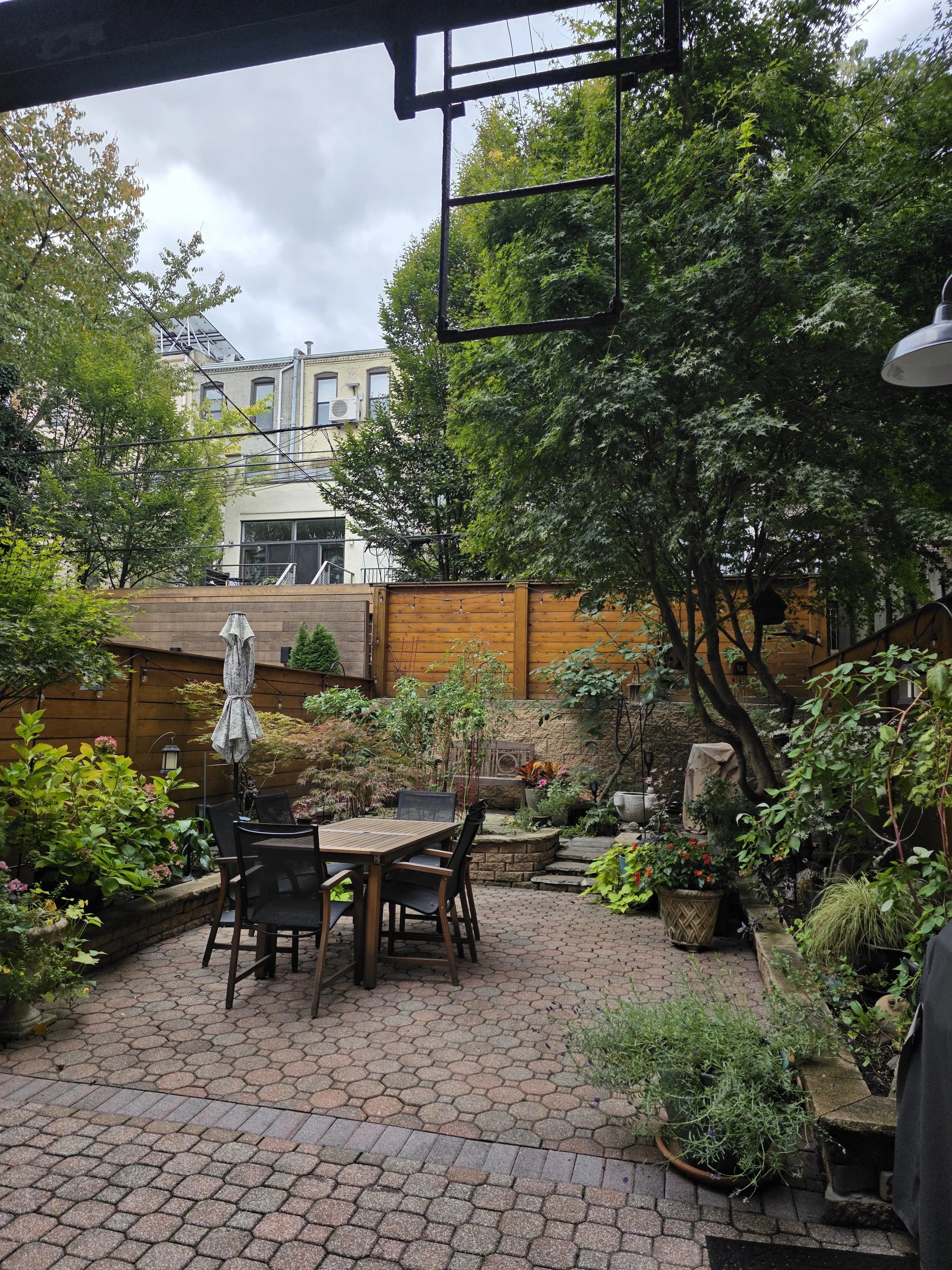 A backyard patio with a round wooden table, six black chairs, an umbrella, potted plants, trees, and a wooden fence, with buildings and trees in the background and an overcast sky.