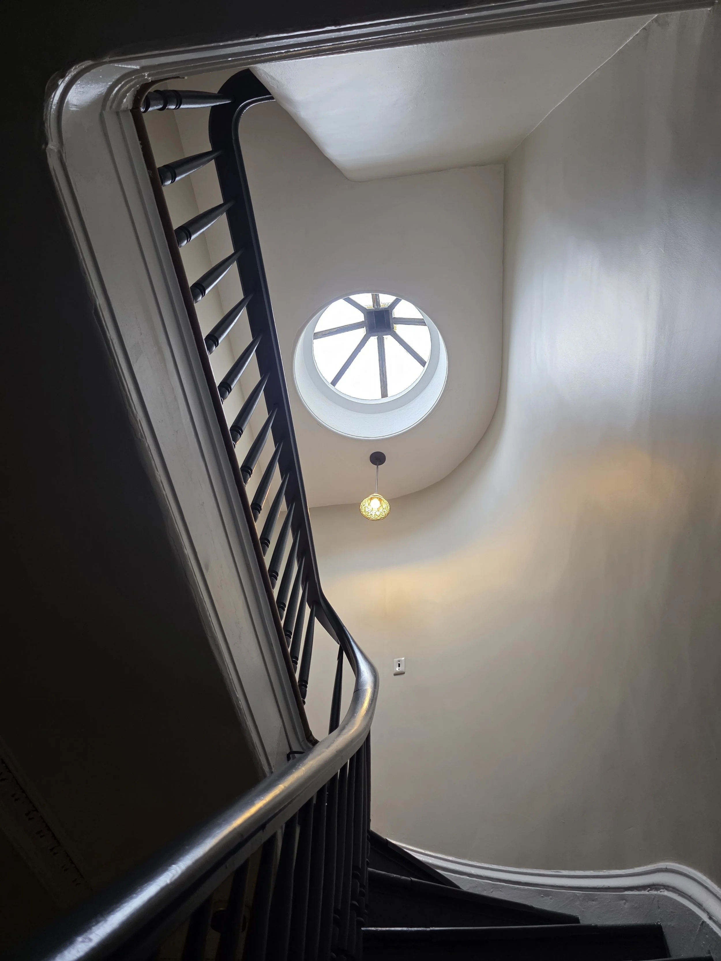 View of an indoor staircase looking up toward a circular skylight with a black frame, with a hanging light fixture below the skylight.