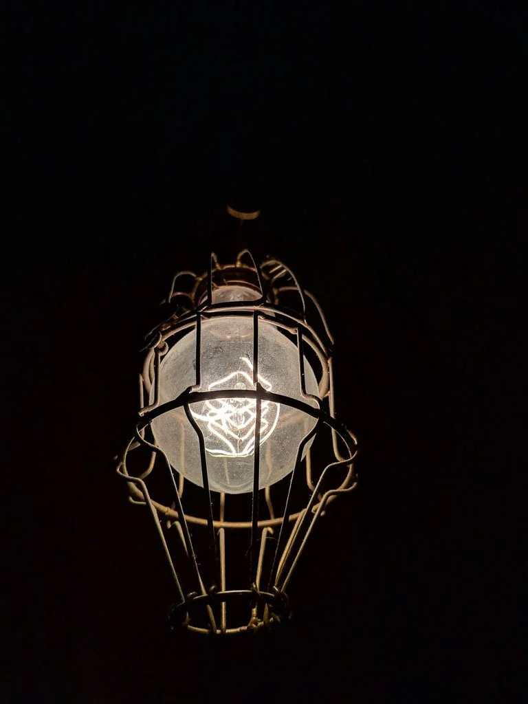 A single lightbulb enclosed in a metal cage glows against a dark background, creating a minimal and moody composition.