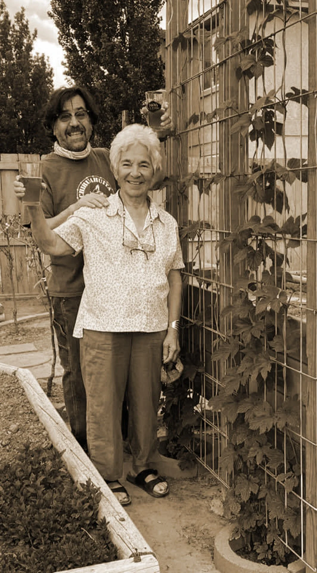 Two people, an older woman and a man, standing outdoors in a garden with a trellis with vines, holding up glasses of beverage and smiling at the camera.