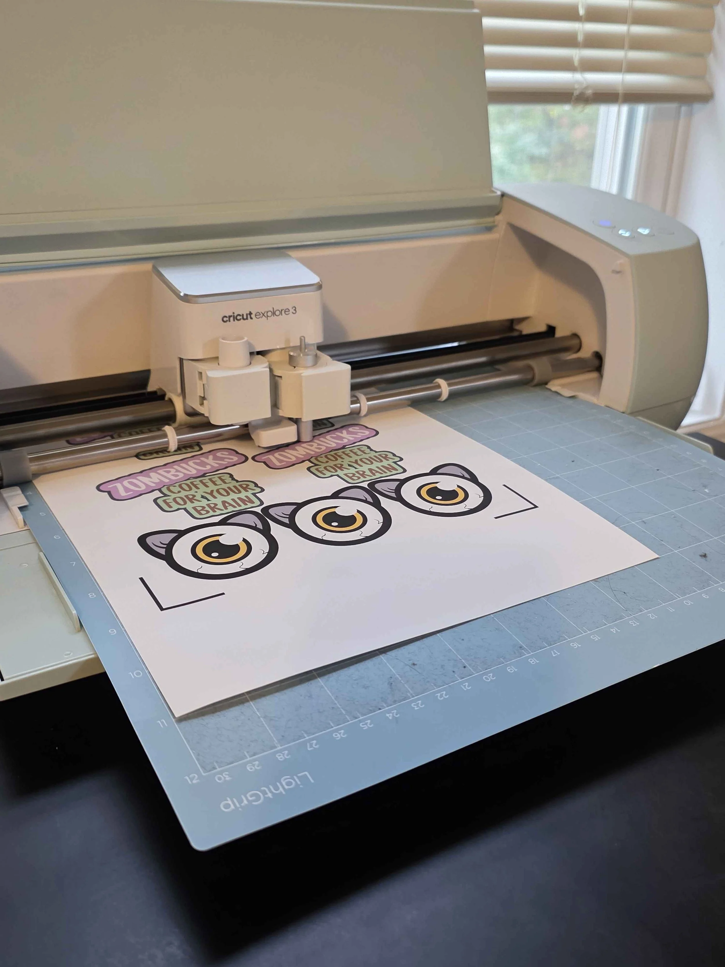 Cricut Explore 3 cutting machine cutting a paper with cartoon cat eyes and slogans 'Zombies' and 'Coffee for your brain'.