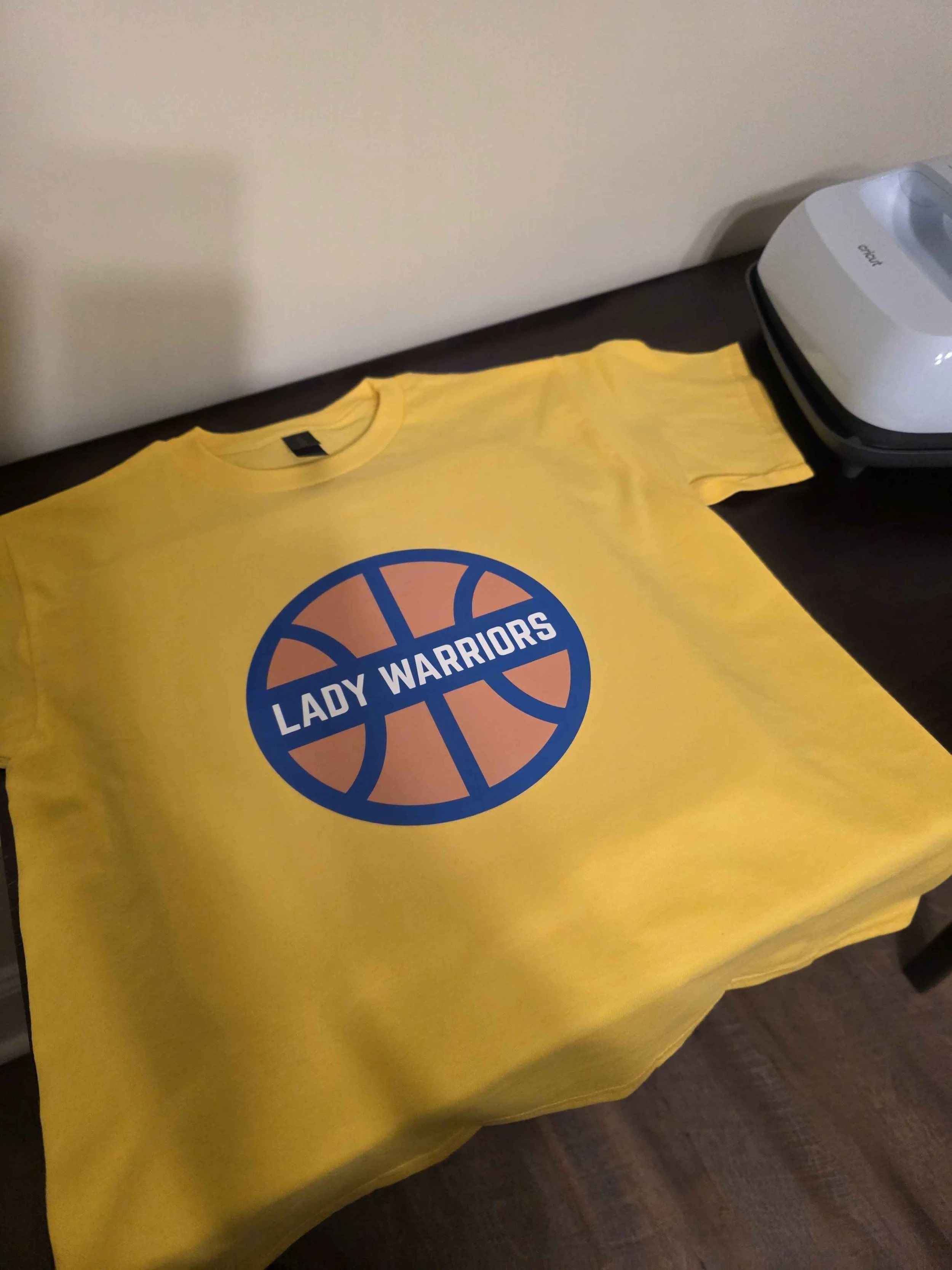 Yellow T-shirt with a blue basketball logo and the words "Lady Warriors" printed on it, placed on a dark table next to a white household device.