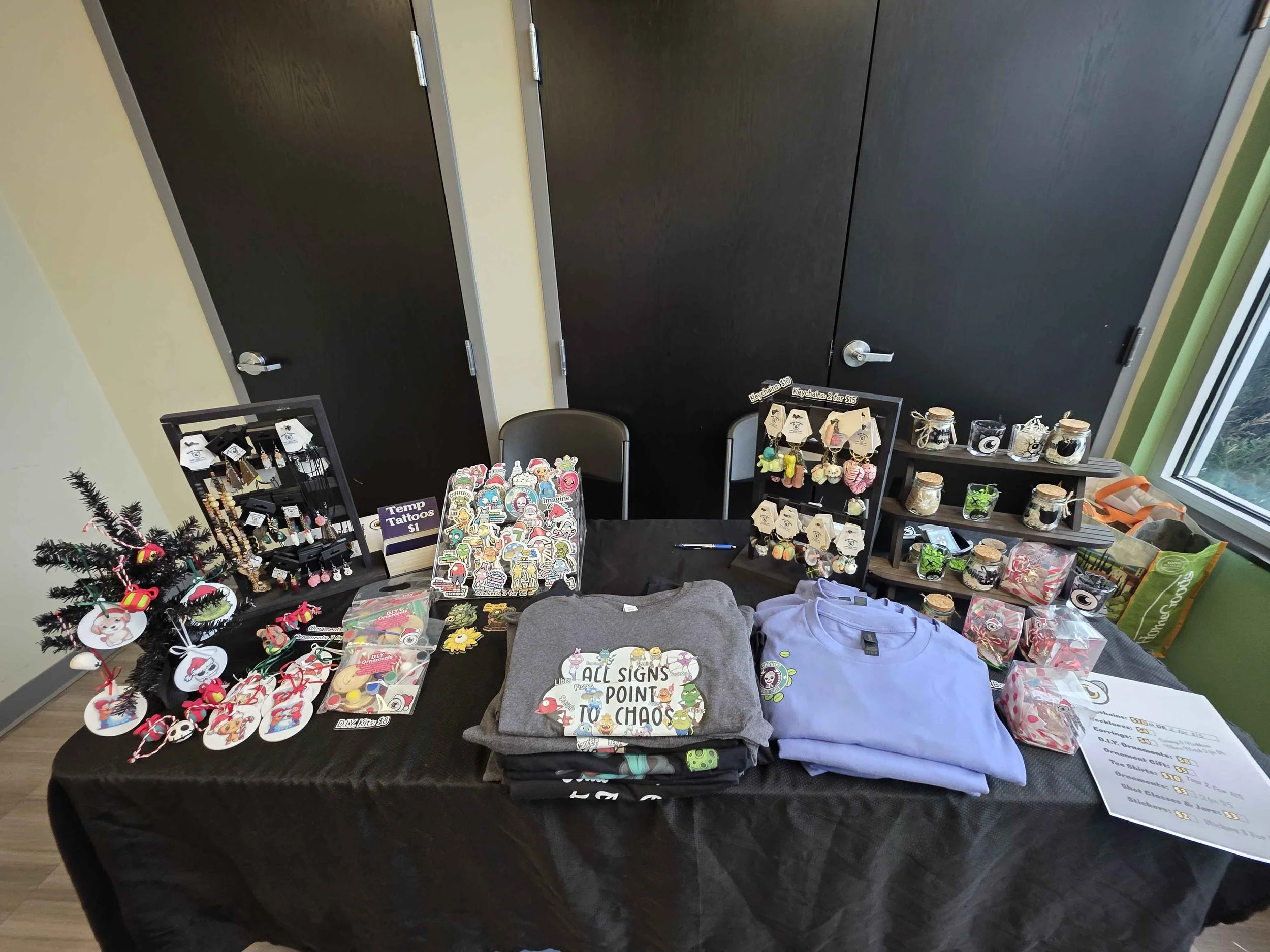 Table with various handmade gifts for sale, including jewelry, keychains, pins, ornaments, and small accessories, arranged neatly on display stands and racks at a craft fair.