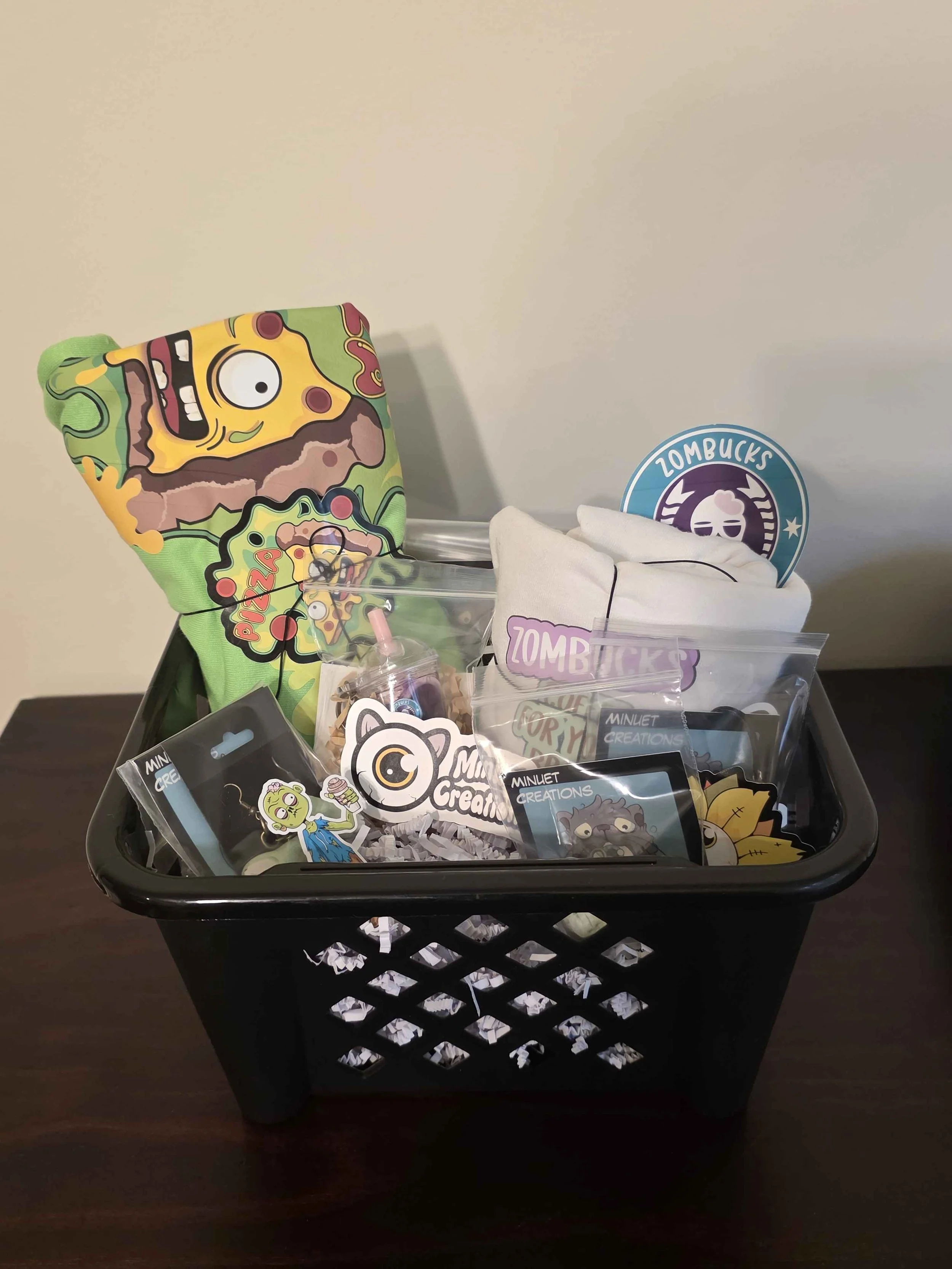 A black plastic basket filled with various colorful items, including a purple zombie-themed pillow, a Zombucks sticker, small packaged accessories, and other zombie-themed merchandise, all arranged on a wooden surface against a plain white wall.