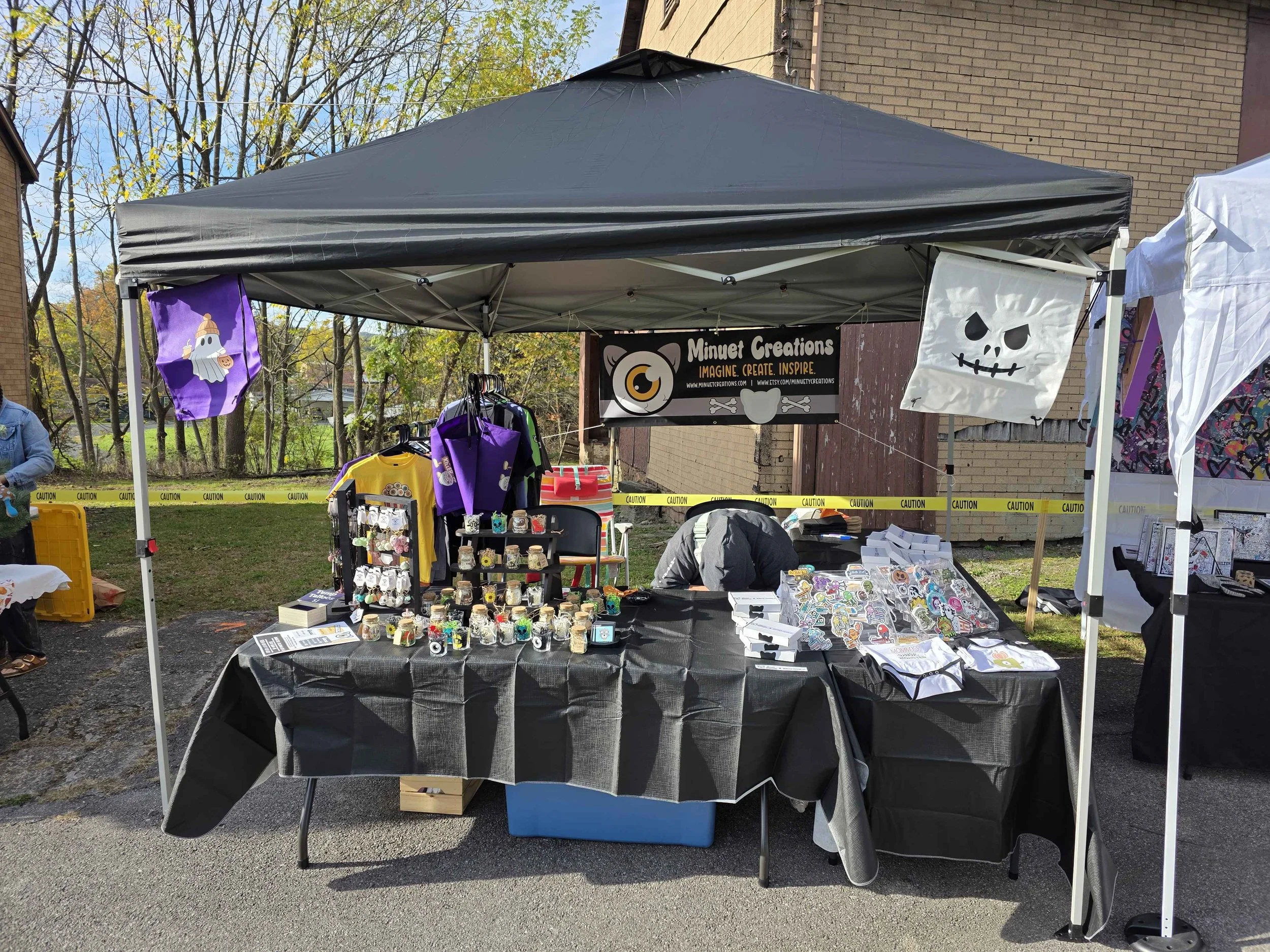 An outdoor vendor stall with a black canopy selling various colorful handmade crafts and stickers, with a banner reading 'Minnert Creations.'