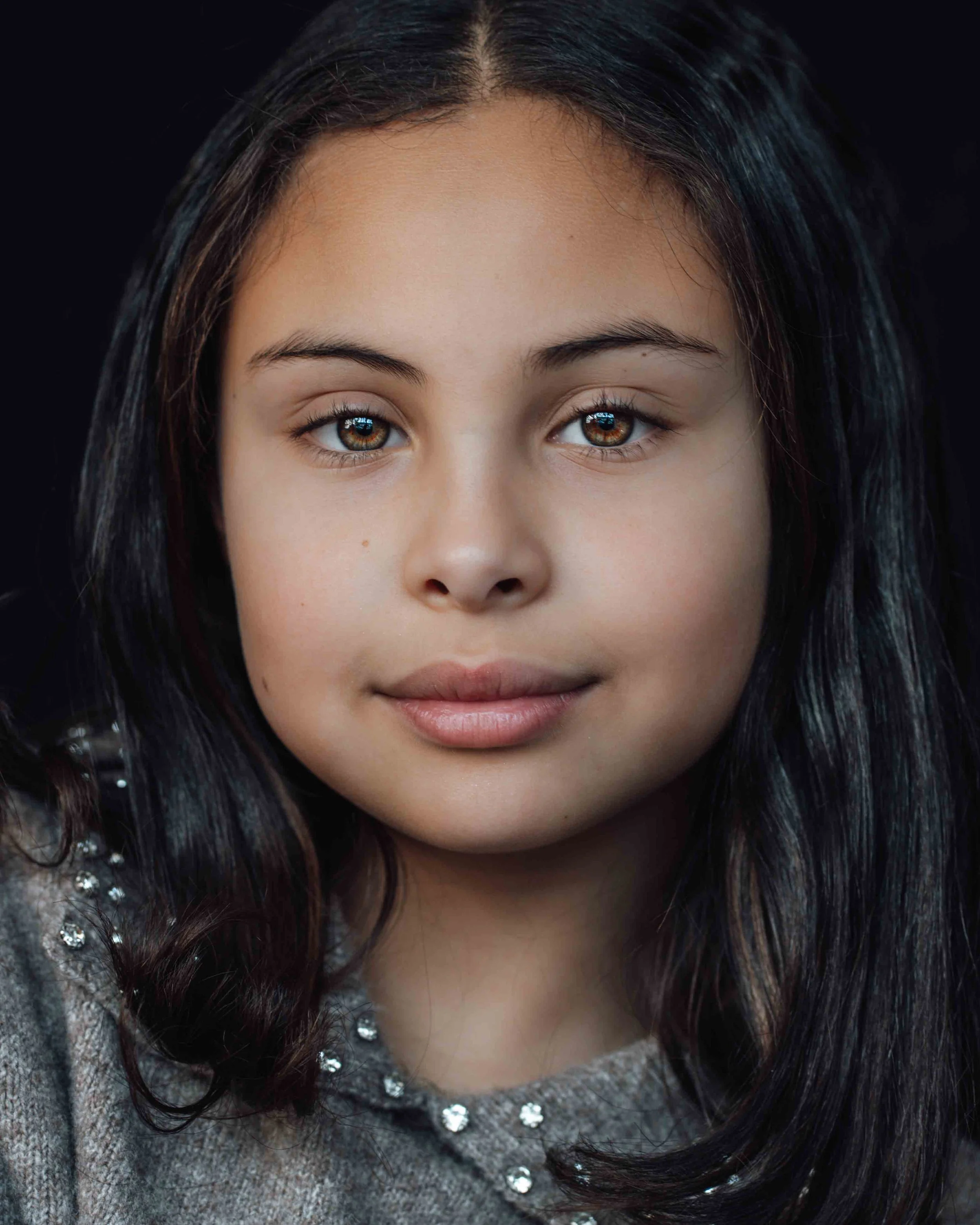 Close-up of a young girl with brown eyes and long dark hair, wearing a grey sweater with small shiny embellishments.