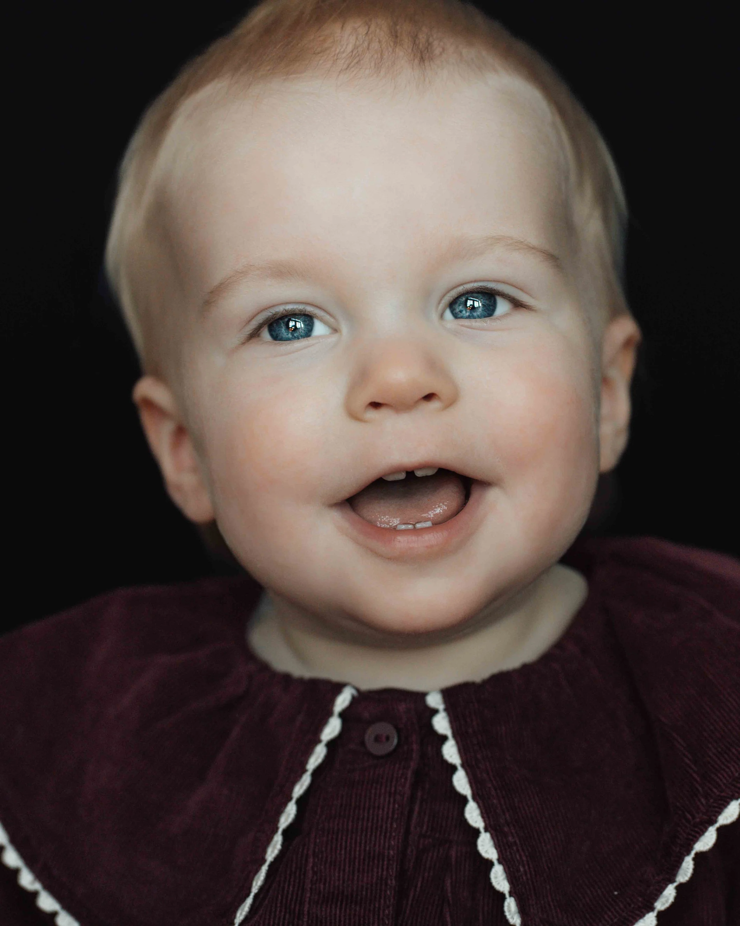 Close-up of a smiling baby with blue eyes, light brown hair, wearing a maroon shirt with white lace trim, against a black background.