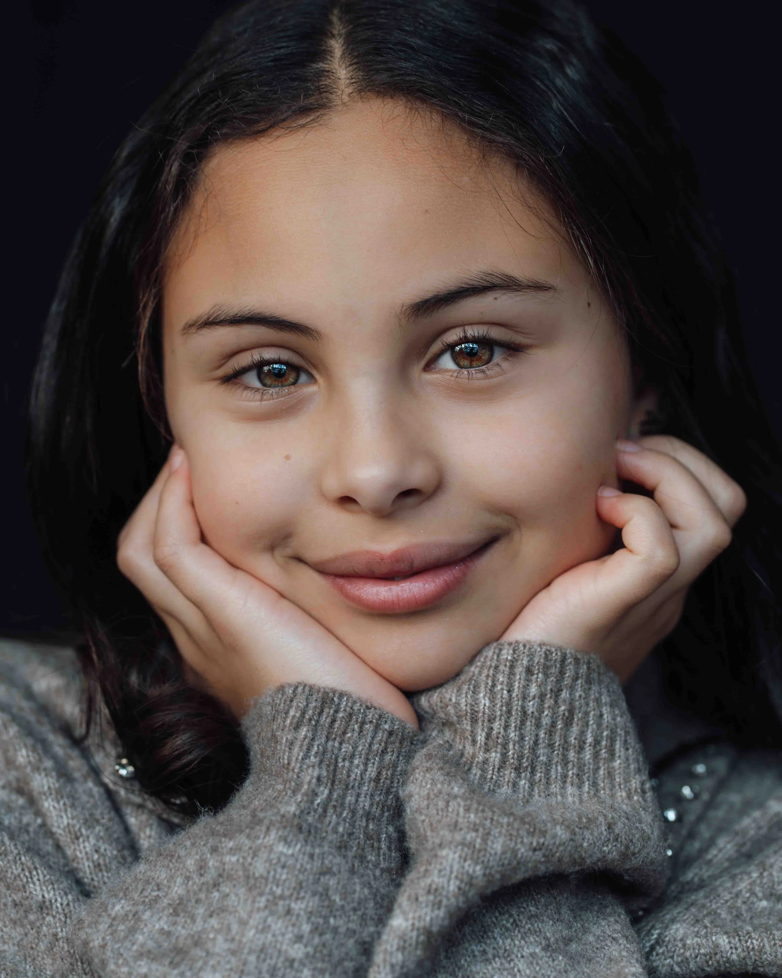 Close-up portrait of a young girl with dark hair, smiling gently, hands resting on her cheeks, wearing a grey sweater, against a black background.