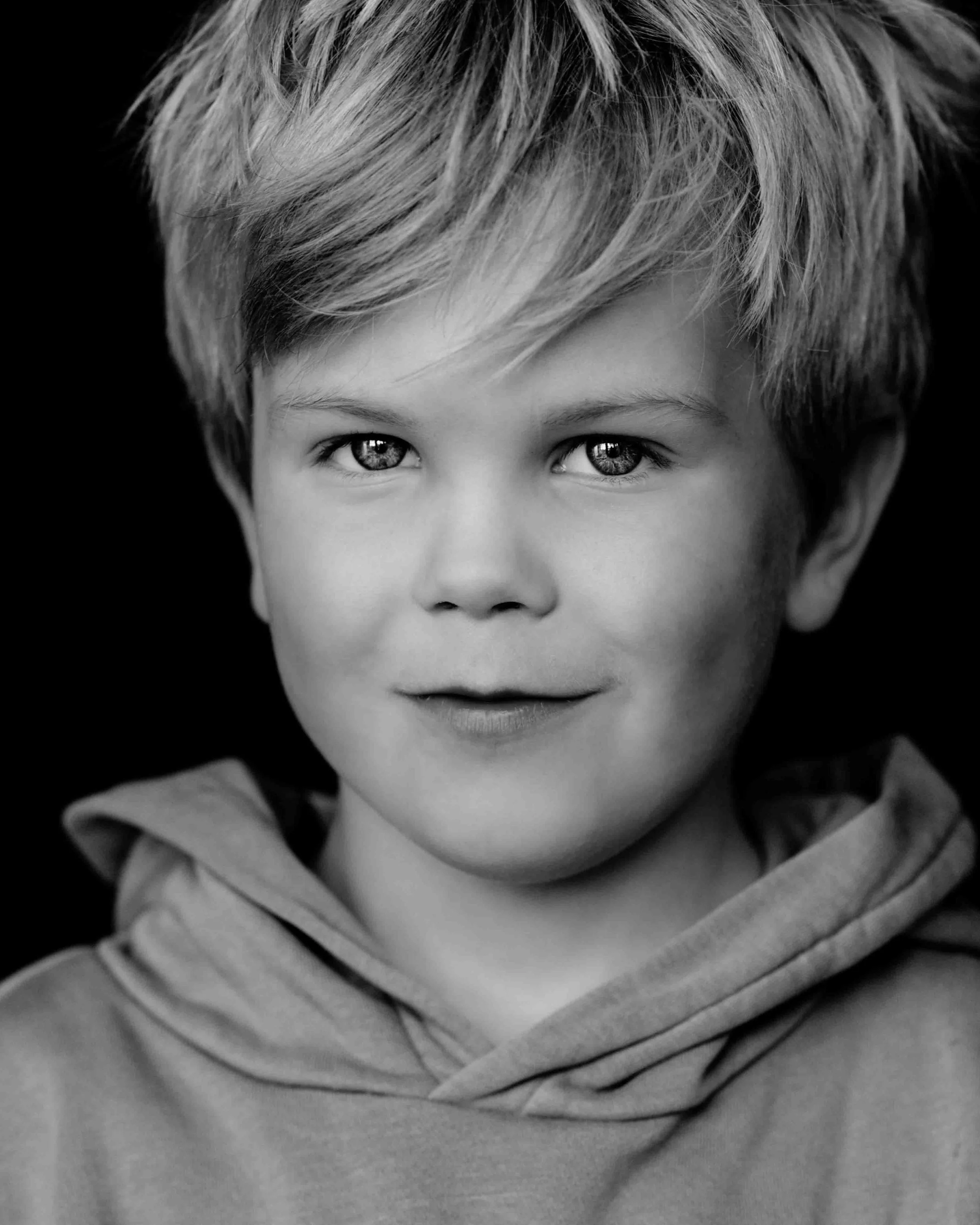 Black and white portrait of a young boy with light-colored hair, wearing a hoodie, looking directly at the camera.