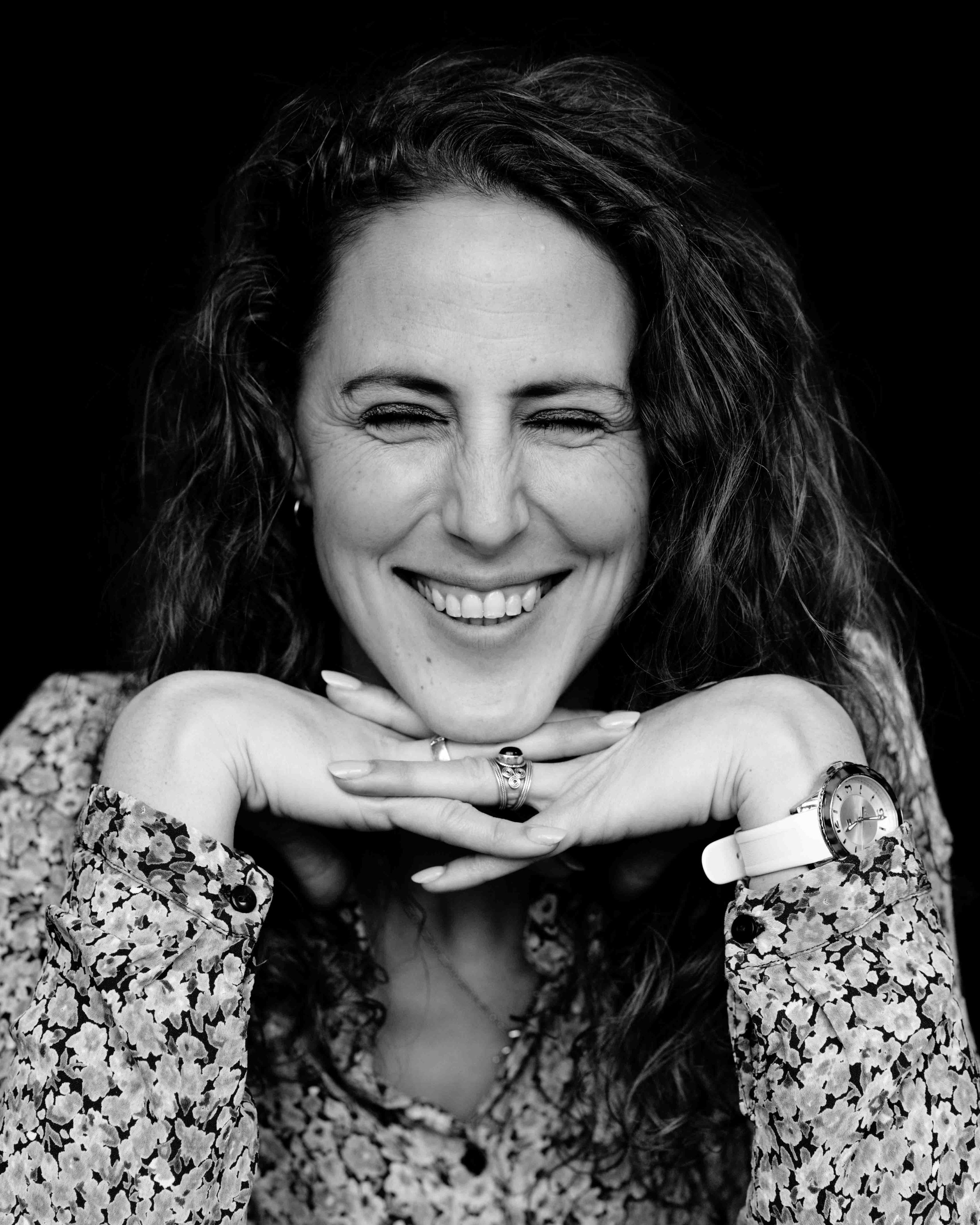 Black and white portrait of a woman smiling with eyes closed, hands resting under her chin, wearing a patterned long-sleeve top, watch, rings, and earrings, against a dark background.